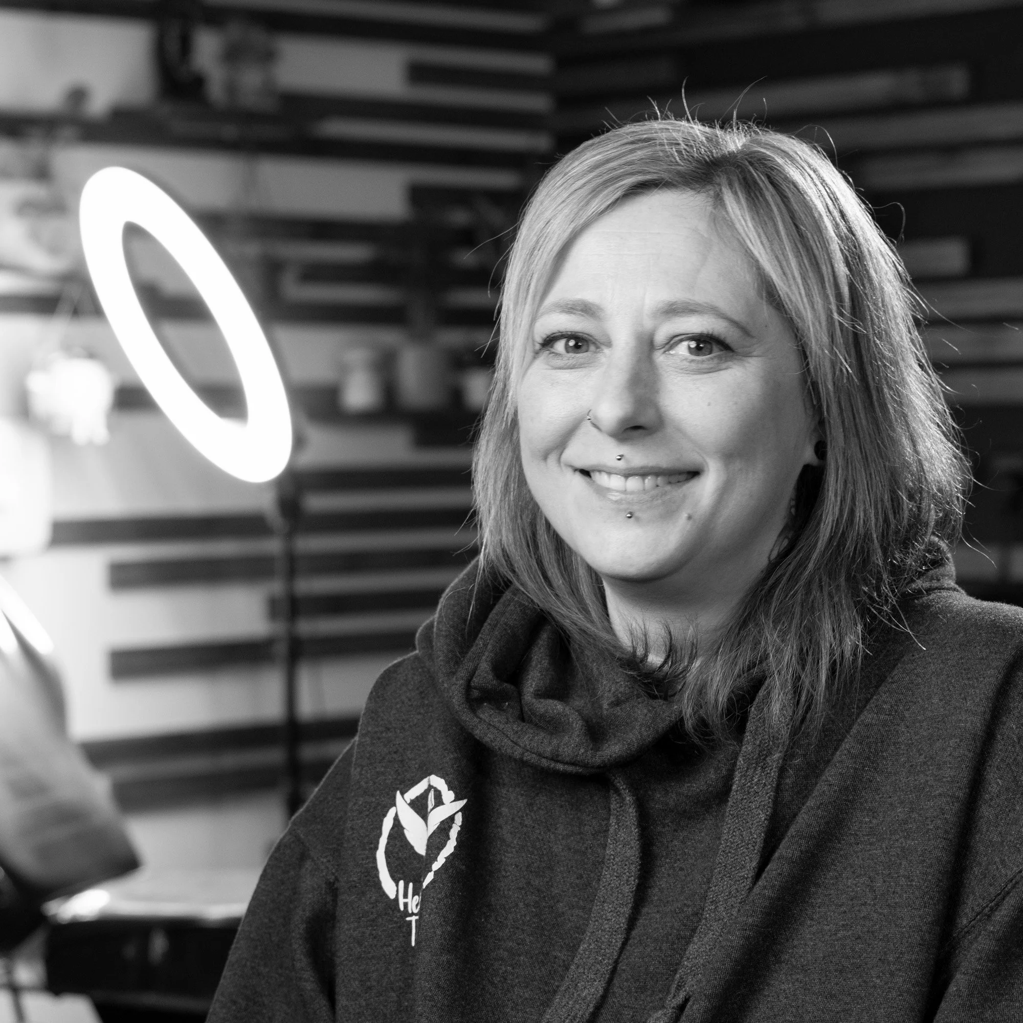 A close-up black-and-white photograph of a woman with light-colored hair, smiling, wearing a dark hoodie with a partially visible logo on the chest, standing in front of a background with wooden slats and a ring light.