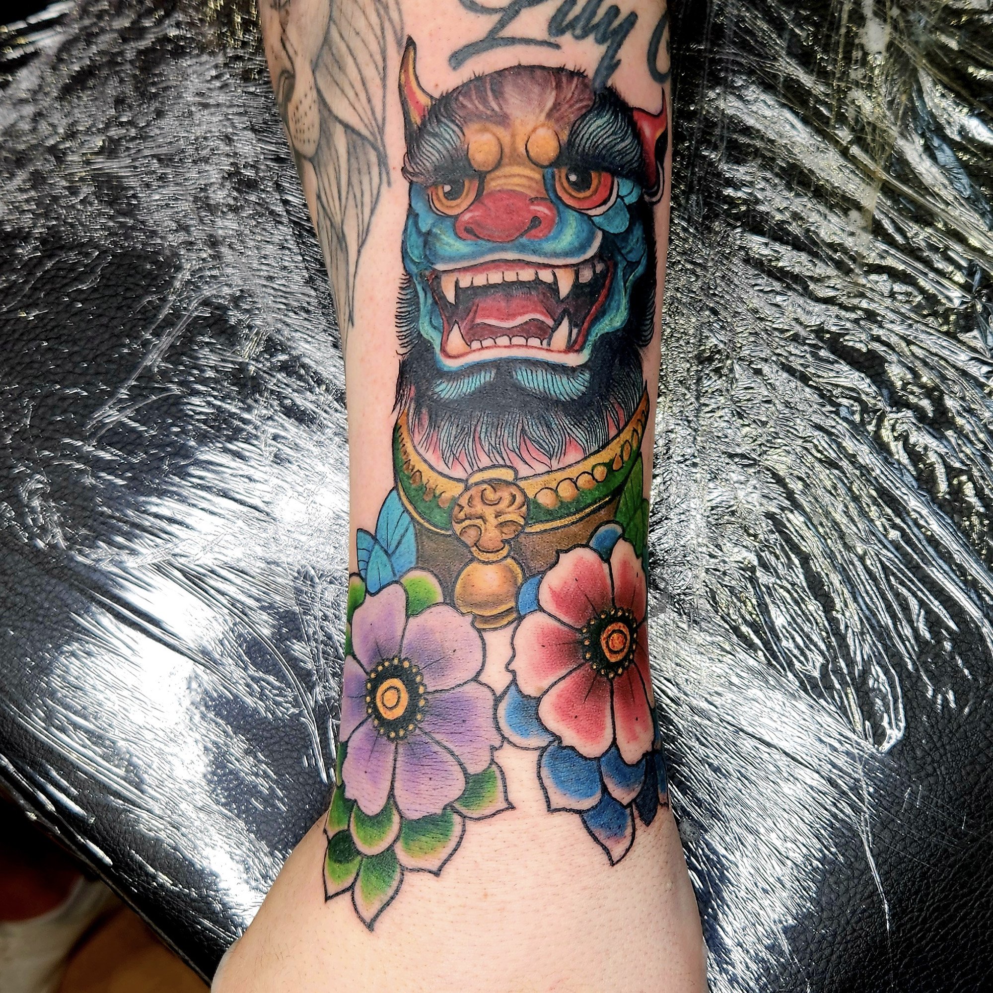 Colorful tattoo of a traditional-style demon or oni face on a person's arm, featuring horns, sharp teeth, and vibrant floral elements surrounding it.
