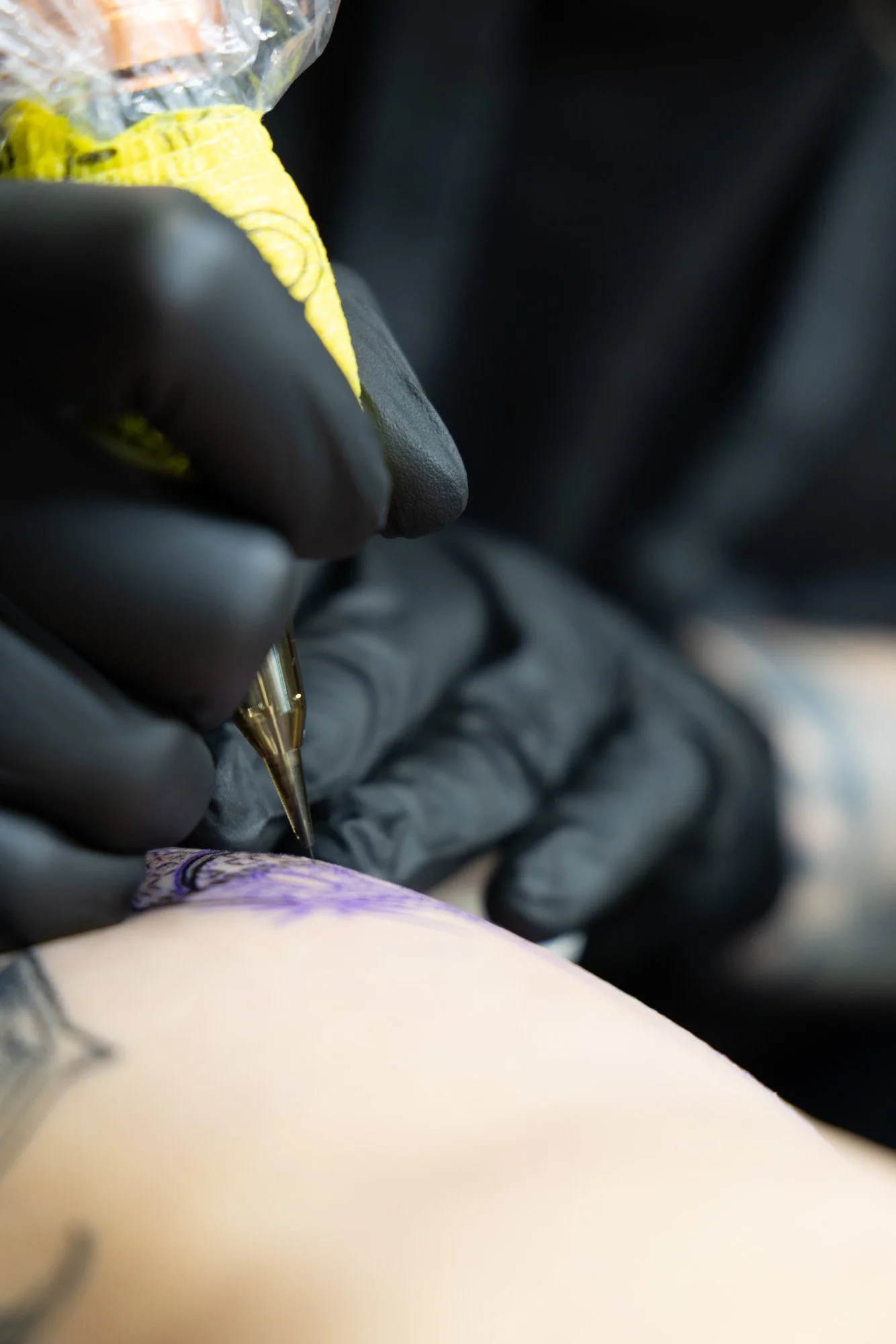 A tattoo artist is inking a design onto a person's skin. The artist is wearing black gloves and using a tattoo machine.