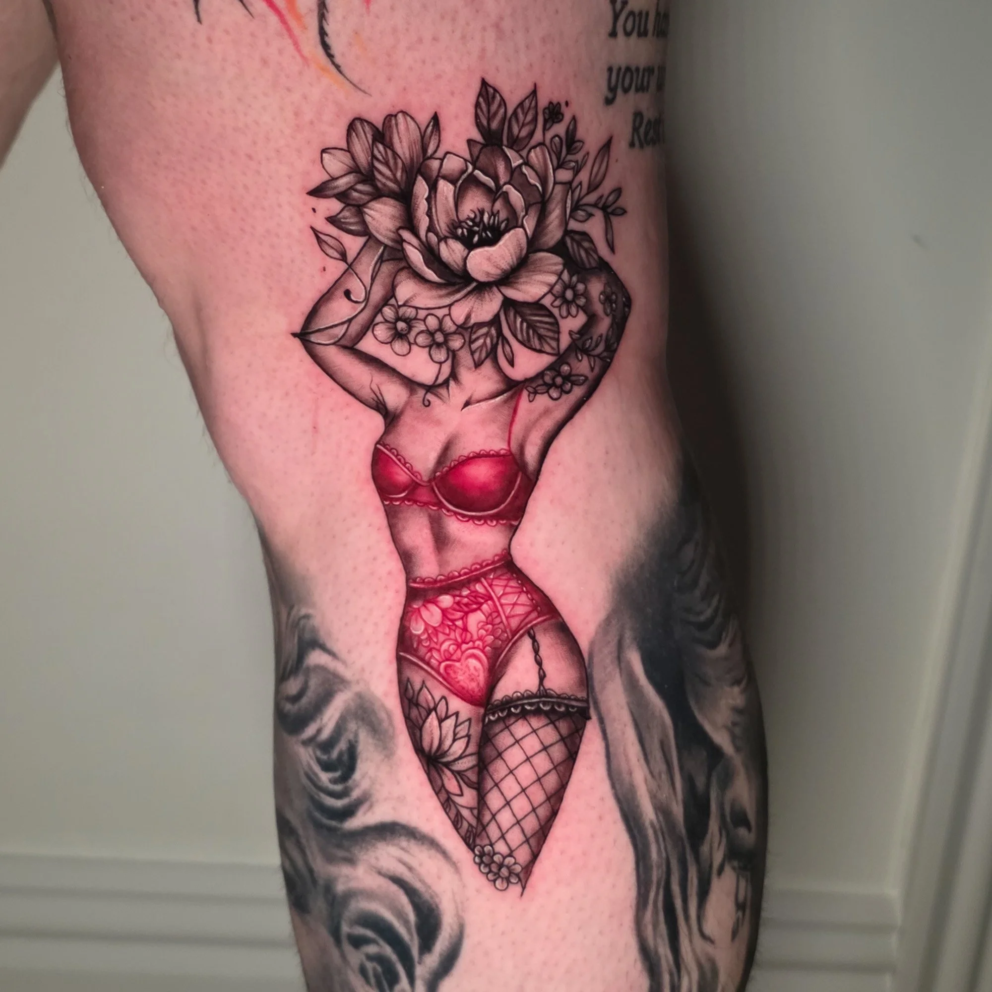 Black and gray floral tattoo on arm with a woman figure wearing red lingerie, tattoo includes roses, leaves, and other flowers, along with some part of text.