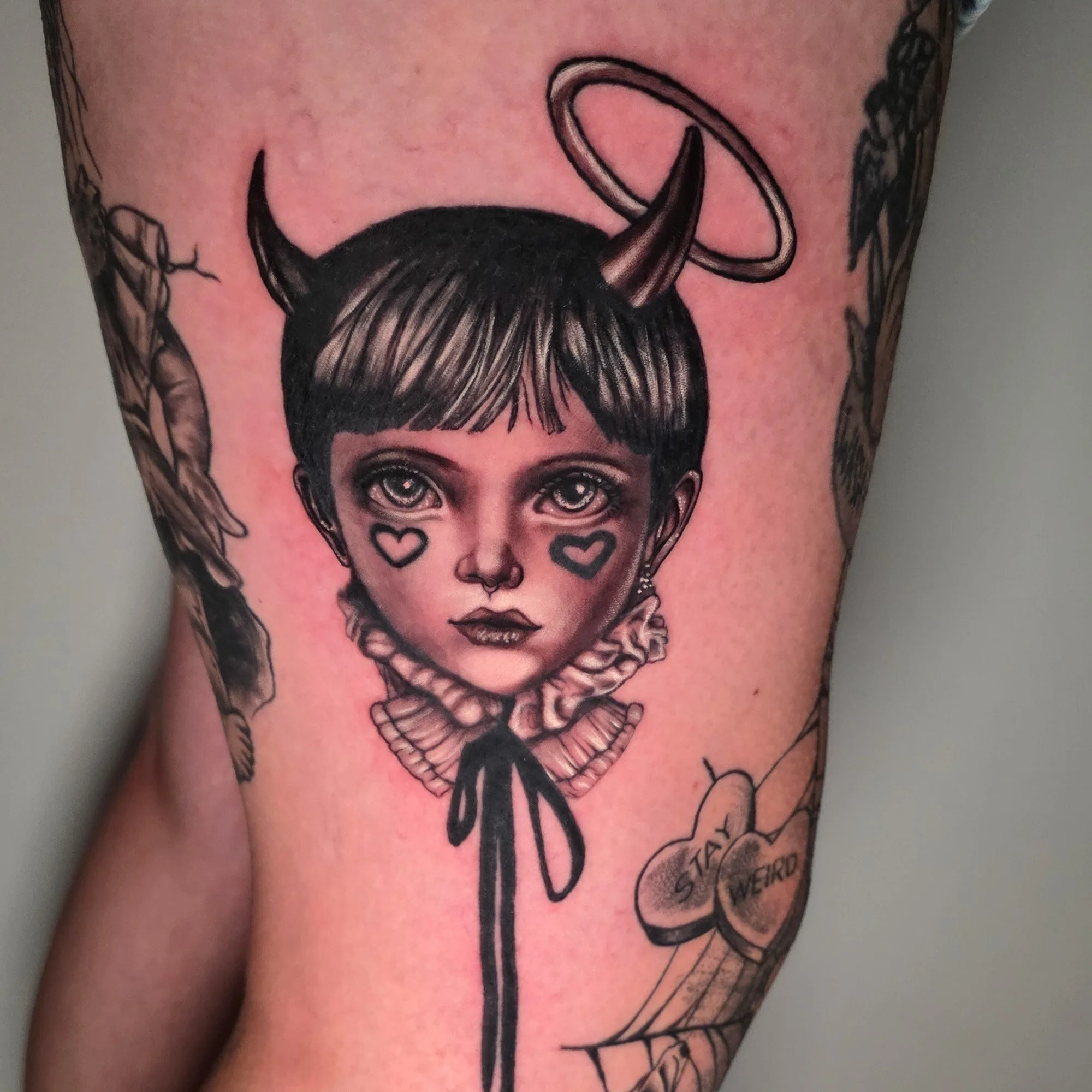 Close-up of a tattoo on skin depicting a stylized woman's face with dark hair, big eyes, and heart-shaped cheek piercings, with additional elements and text surrounding the face.