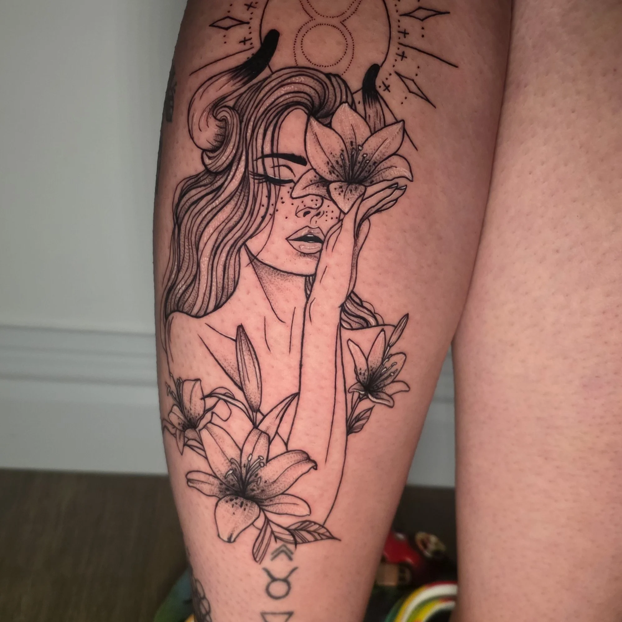 A tattoo on skin featuring a woman's face with long flowing hair, surrounded by large lilies and various geometric and decorative elements.