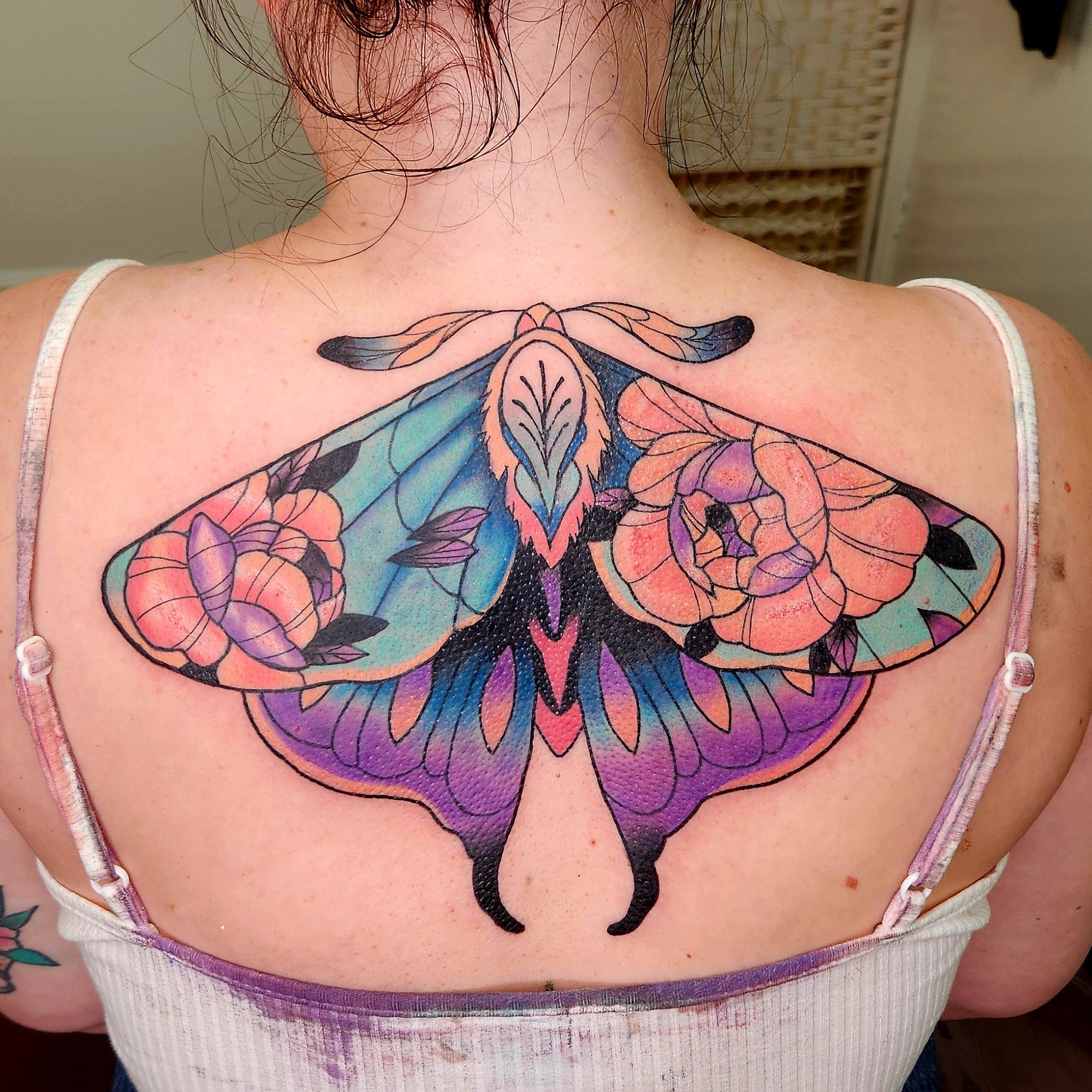 Colorful butterfly tattoo on person's upper back with a background of skin and a white tank top strap visible.