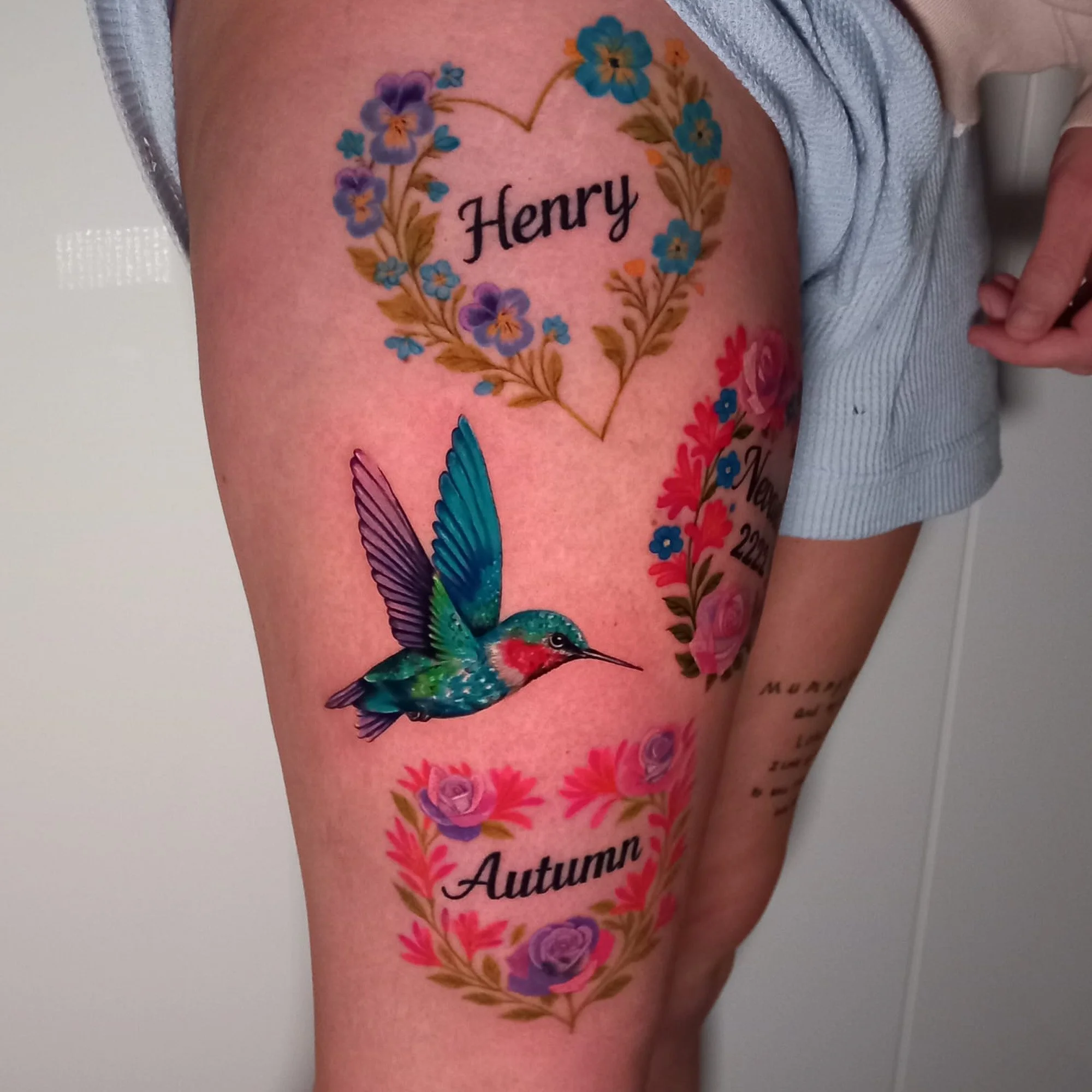 A tattoo on a person's arm featuring a heart shape made of blue and purple flowers and leaves, with the name 'Henry' written inside the heart.