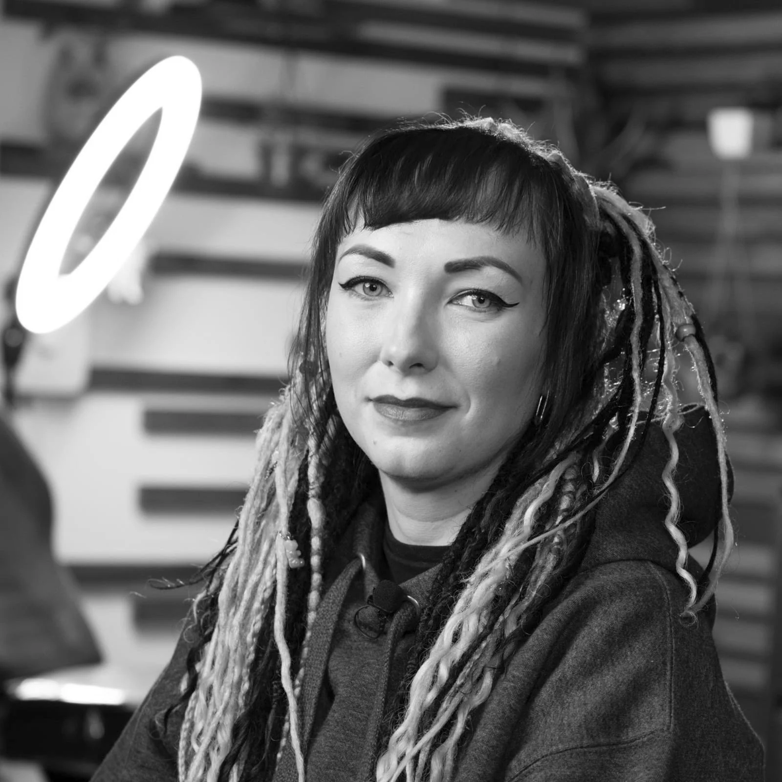 A tattoo artist with short dark hair, light-colored eyes, and a nose ring, wearing a dark hoodie, sitting at a table in an indoor setting with a blurred background.