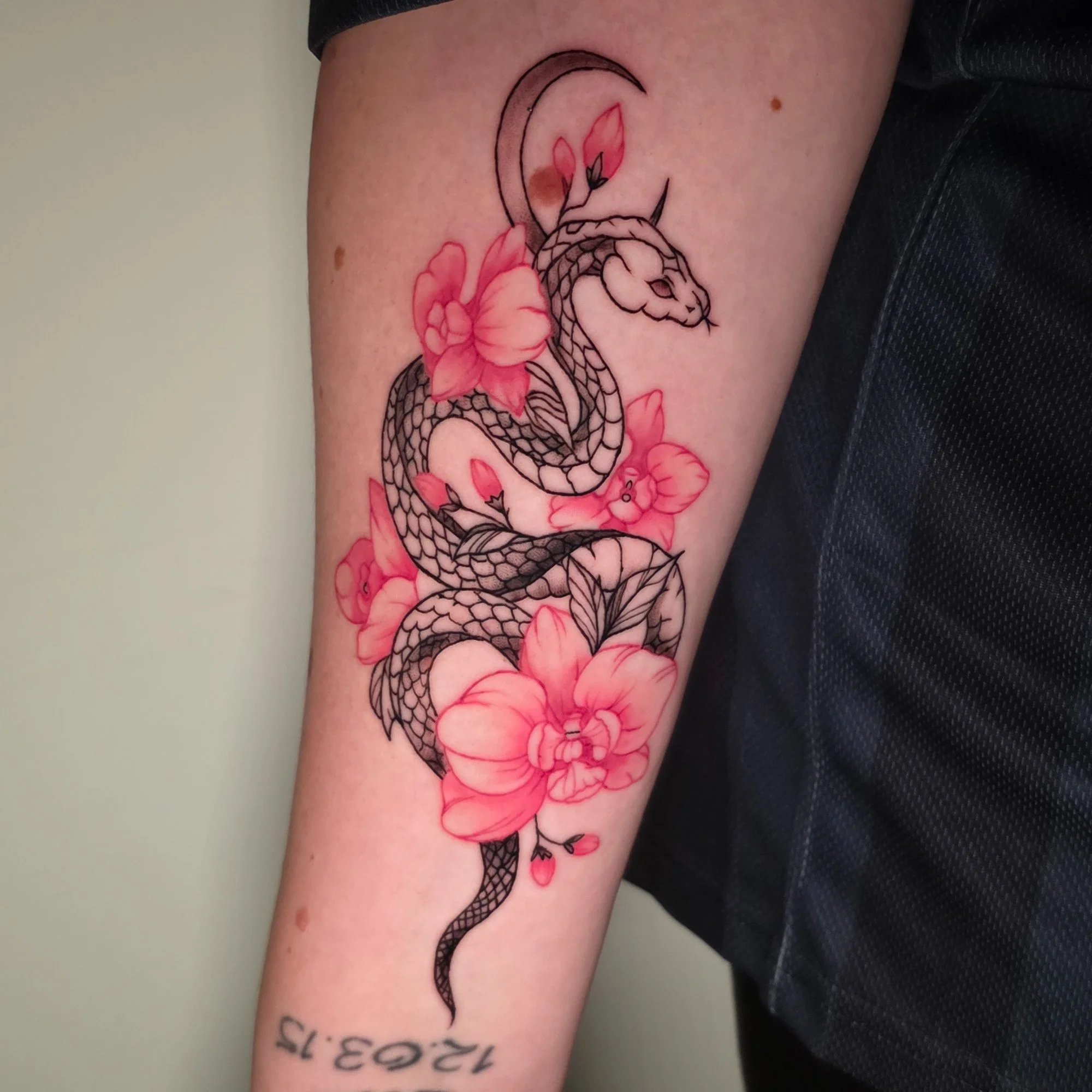 Tattoo of a black outlined snake intertwined with pink flowers on a person's forearm.