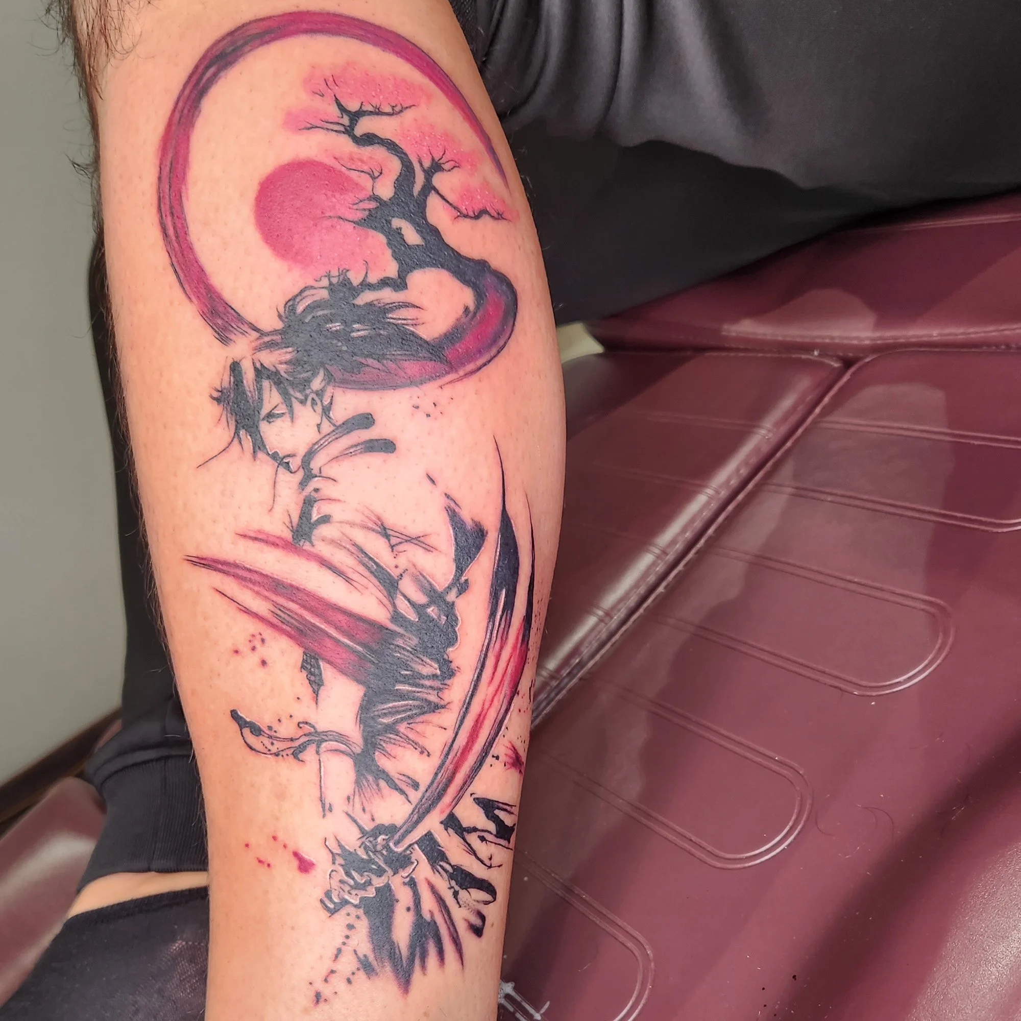 A tattoo of a black tree with many branches inside a pink watercolor circle on a person's upper arm.