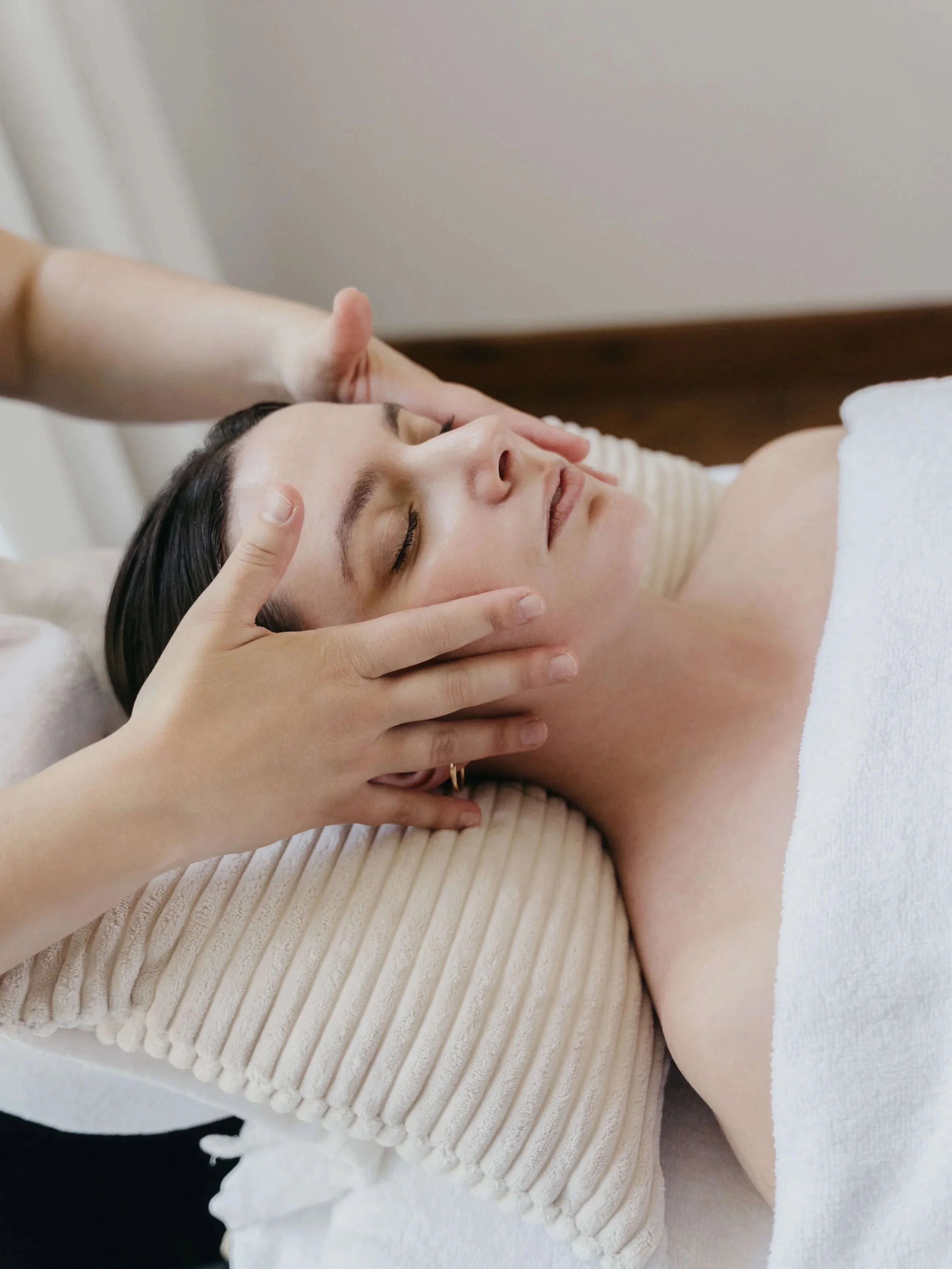 Une femme reçoit un massage facial, couché sur un lit avec une serviette blanche, visage détendu, dans un environnement apaisant.