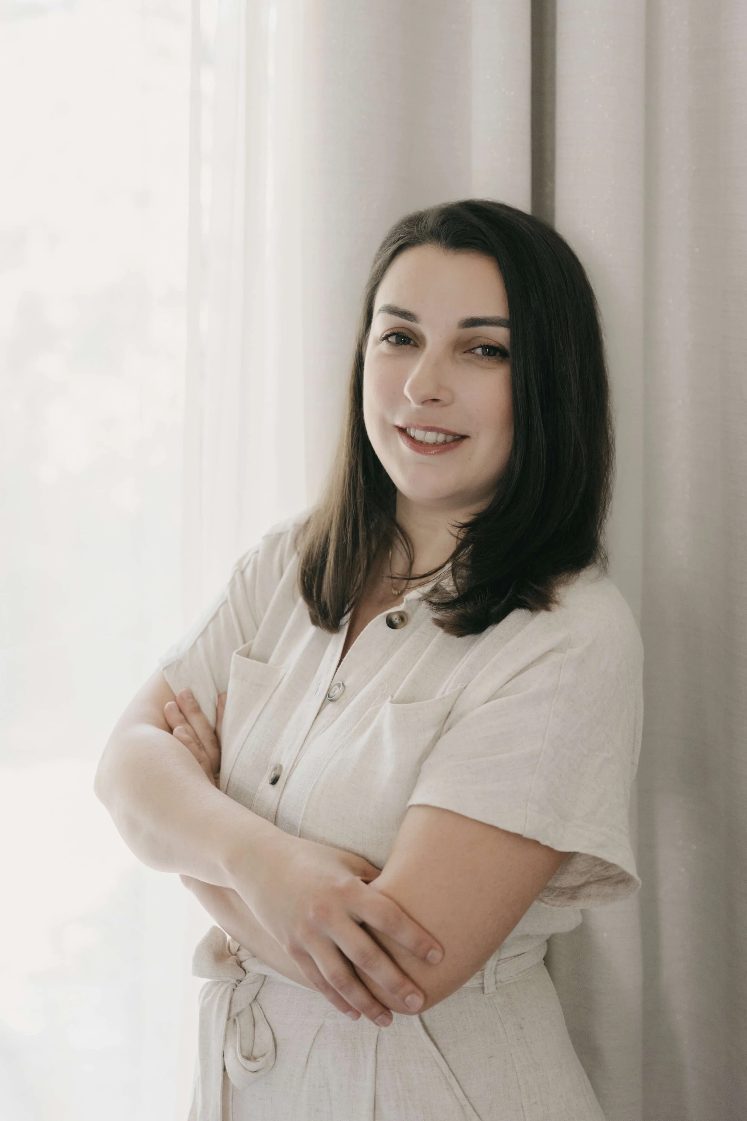 Portrait d'une femme massothérapeute, masseuse, avec des cheveux bruns, souriant, portant une tenue beige, avec des bras croisés, devant un rideau blanc.