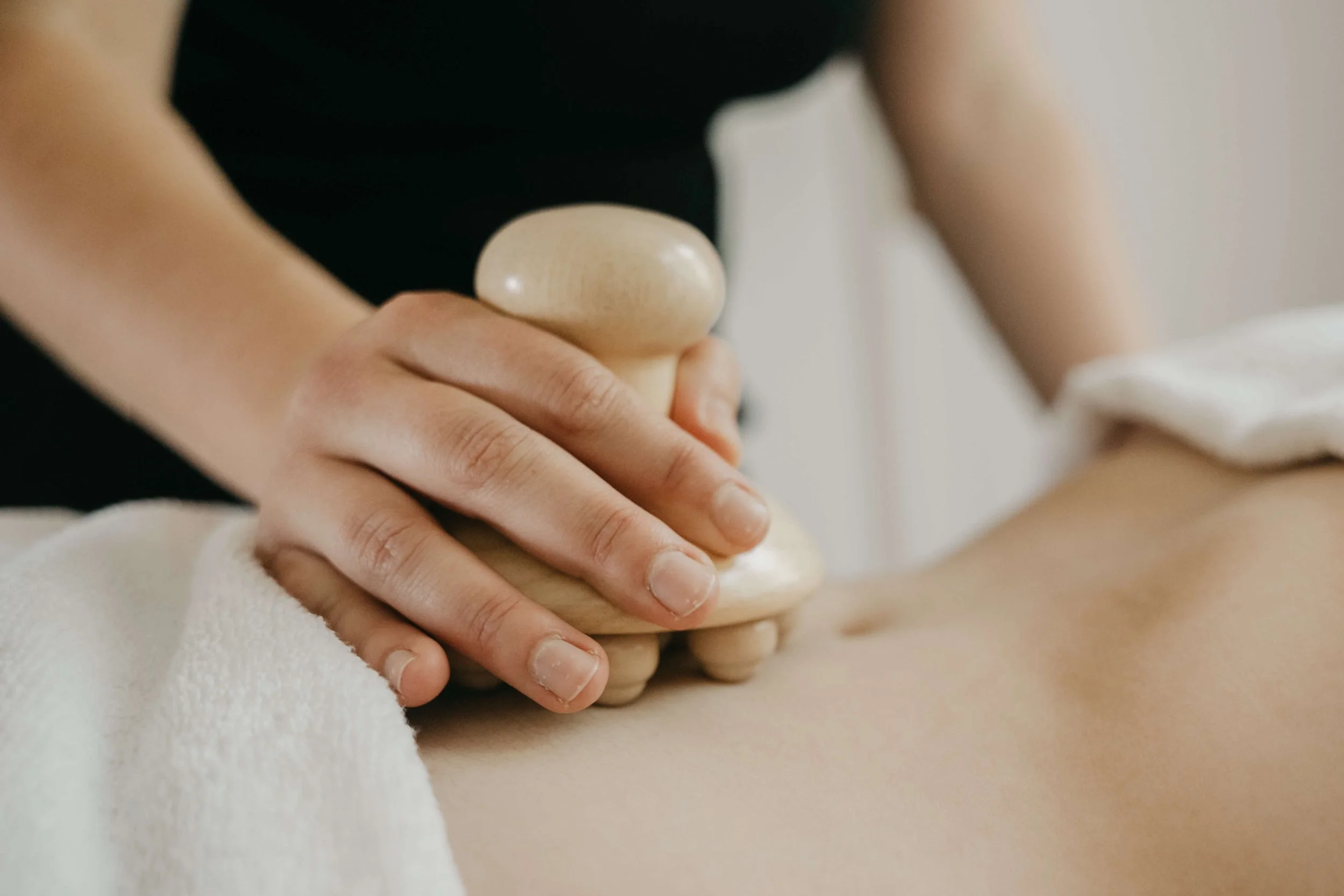 Une personne utilise un rouleau en bois de madérothérapie pour faire un massage sur une autre personne, la main de la personne qui reçoit le massage repose sur du linge en coton blanc.