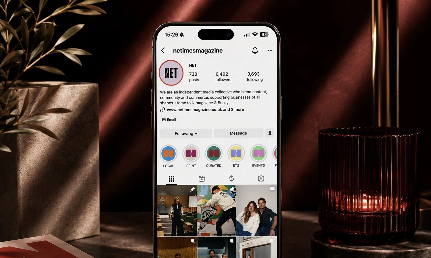 Instagram profile page of netimesmagazine displayed on a smartphone, with a plant in a planter on the left and a red glass candle holder on the right in a dimly lit room.