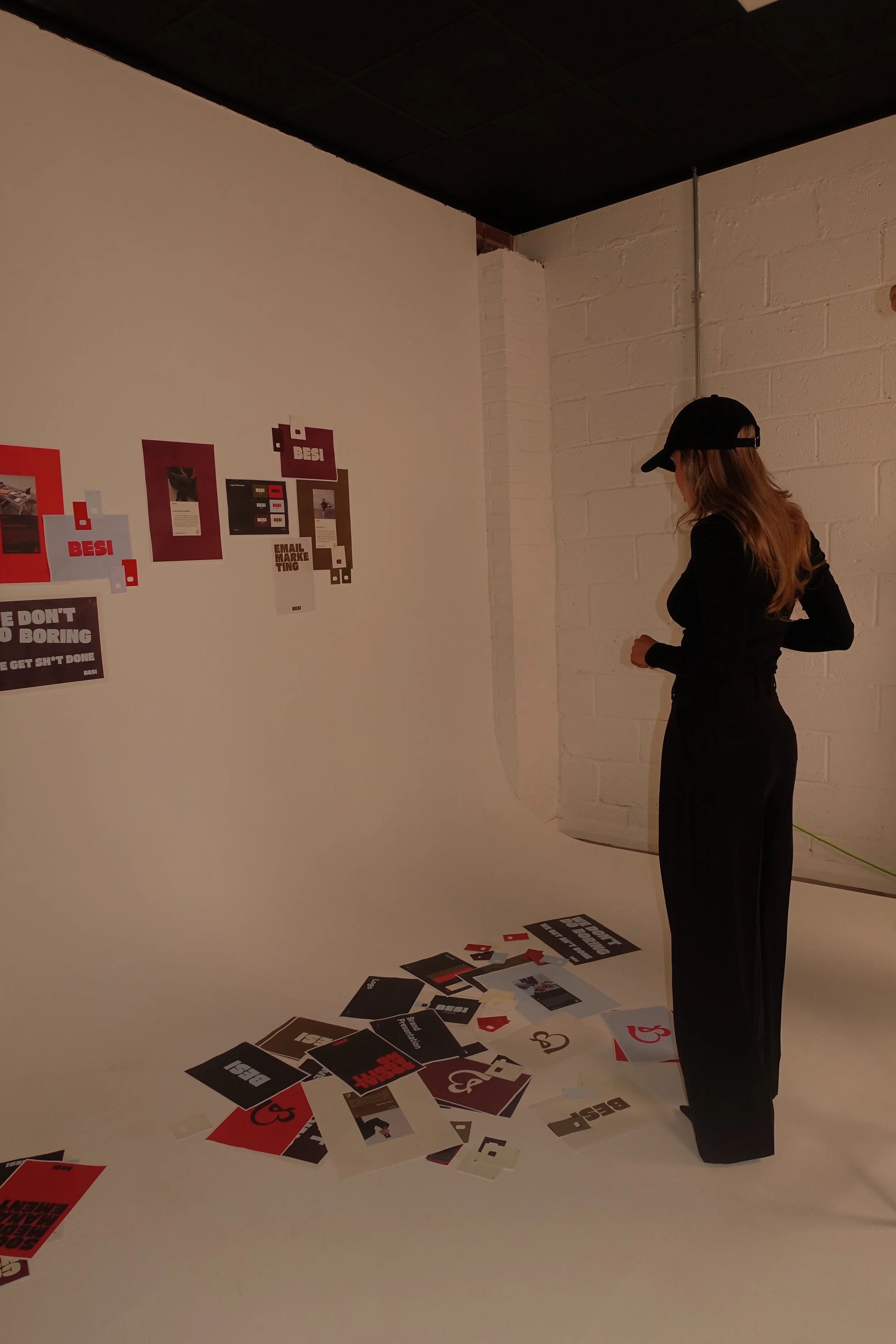 A woman in black clothing and a black cap looking down at scattered black, red, and white papers on the floor and attached to the white wall. The papers feature bold text such as 'BESI Studios', 'Bespoke marketing', and Social media management'.