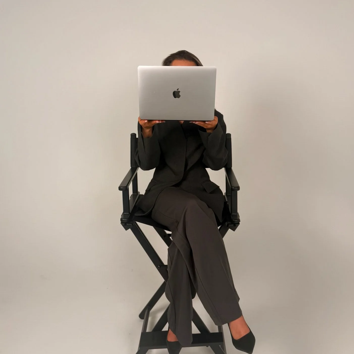A person dressed in a black blazer and grey pants sitting on a director's chair, holding an open silver Apple laptop in front of their face against a plain white wall.