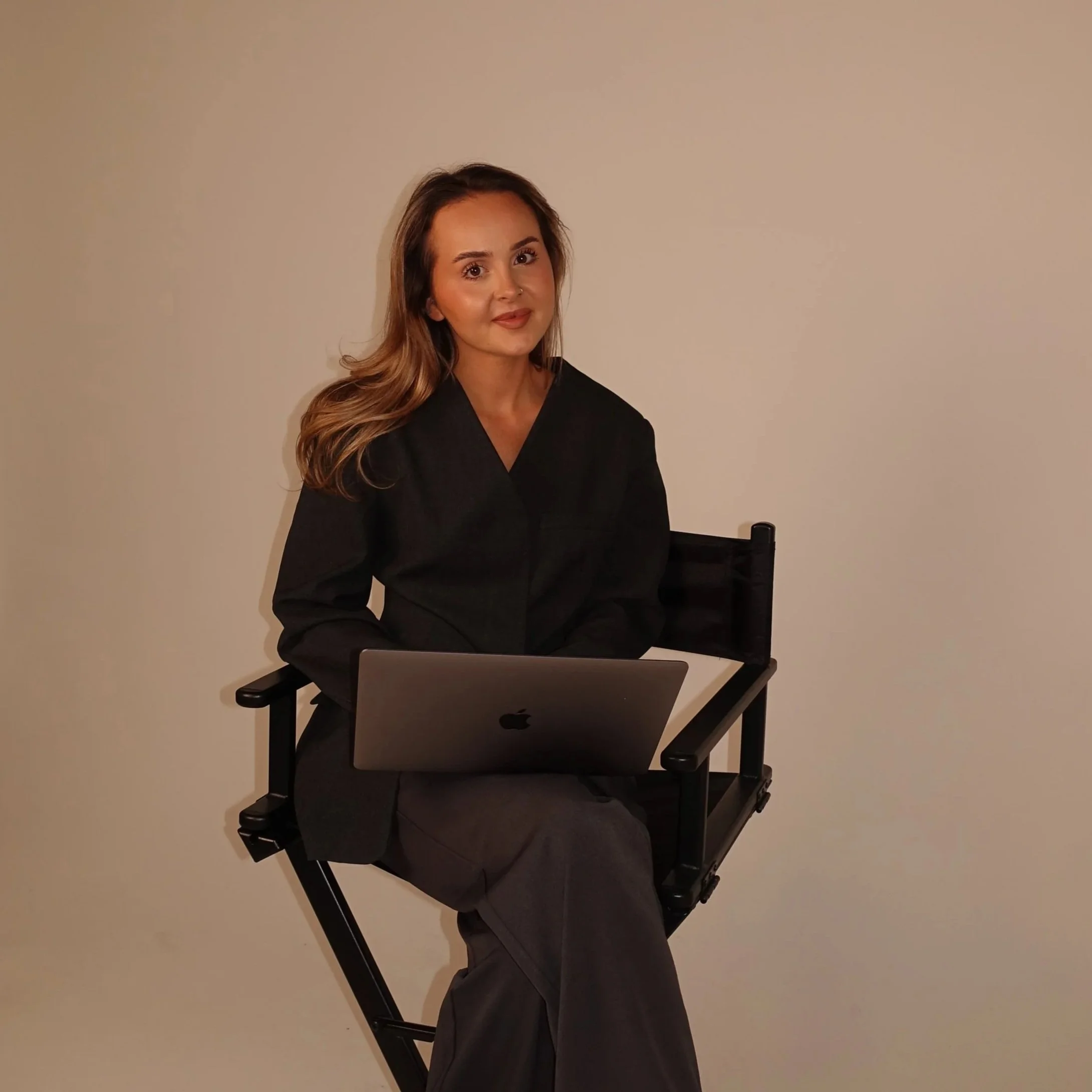 A businesswoman with long wavy hair, wearing a black top, sitting in front of a plain beige wall, looking at the camera. There is a laptop on her knee in front of her.