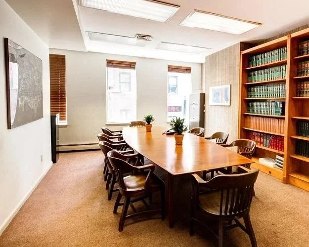 Sala de reunião com mesa de madeira oval e dez cadeiras ao redor, três plantas ao centro, janelas com cortinas de madeira, estantes de livros na parede direita, quadro na parede esquerda, iluminação de teto, carpete bege no chão.