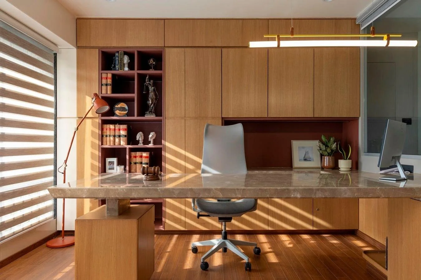 Escritório com mesa de mármore, cadeira cinza, prateleiras de madeira com livros e decorações, luminária de teto amarela, plantas, janela com persianas horizontais.