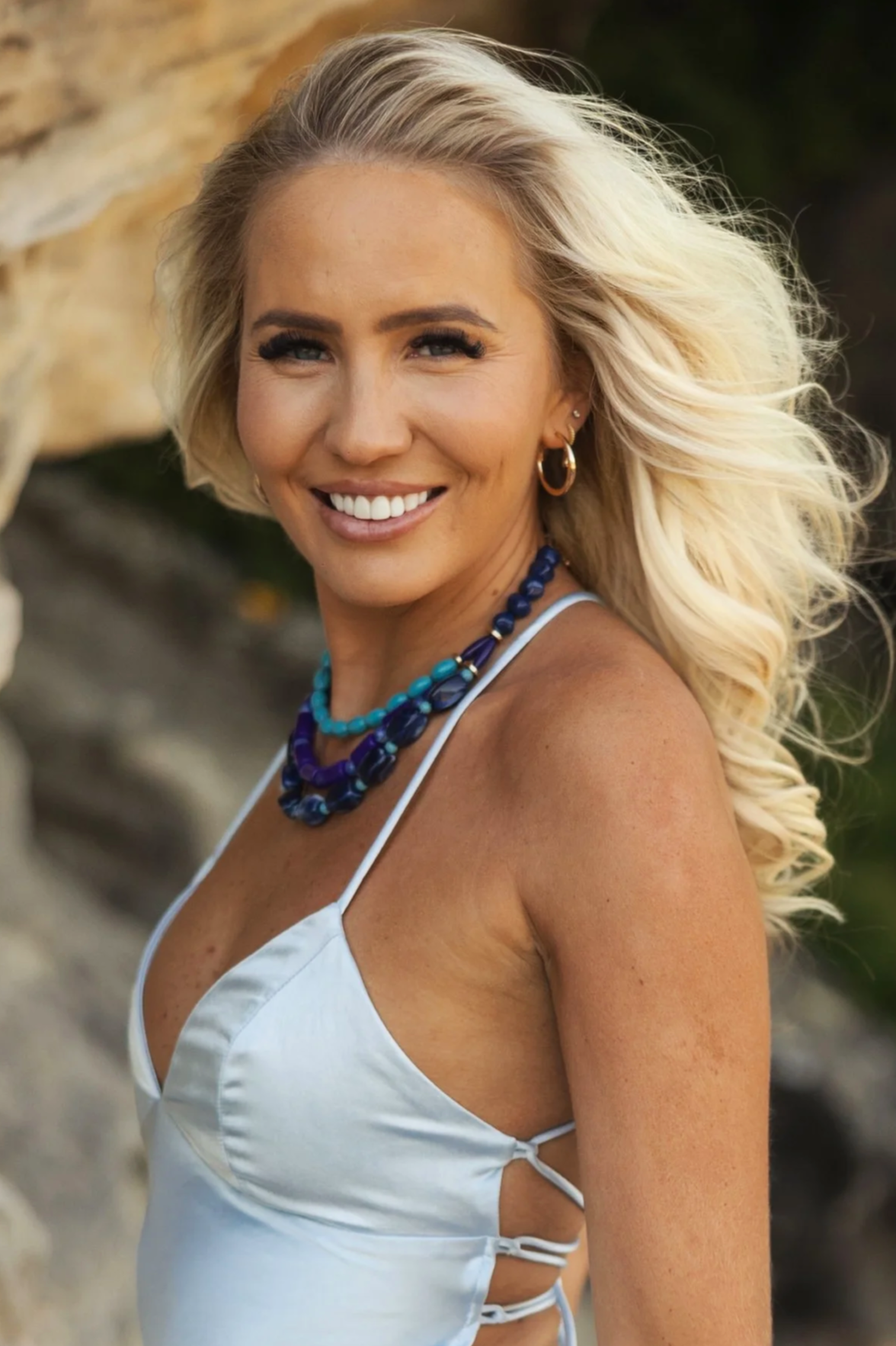 A woman with blonde hair and a white satin dress, smiling outdoors with a blurred rocky background, wearing colorful jewelry.