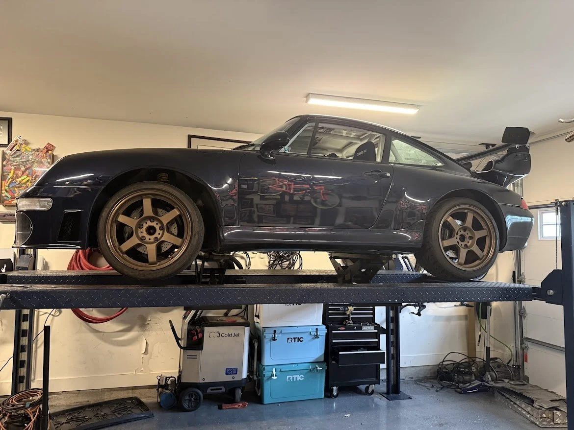 Black sports car with a rear spoiler, brown wheels, and tinted windows on a lift in a garage.