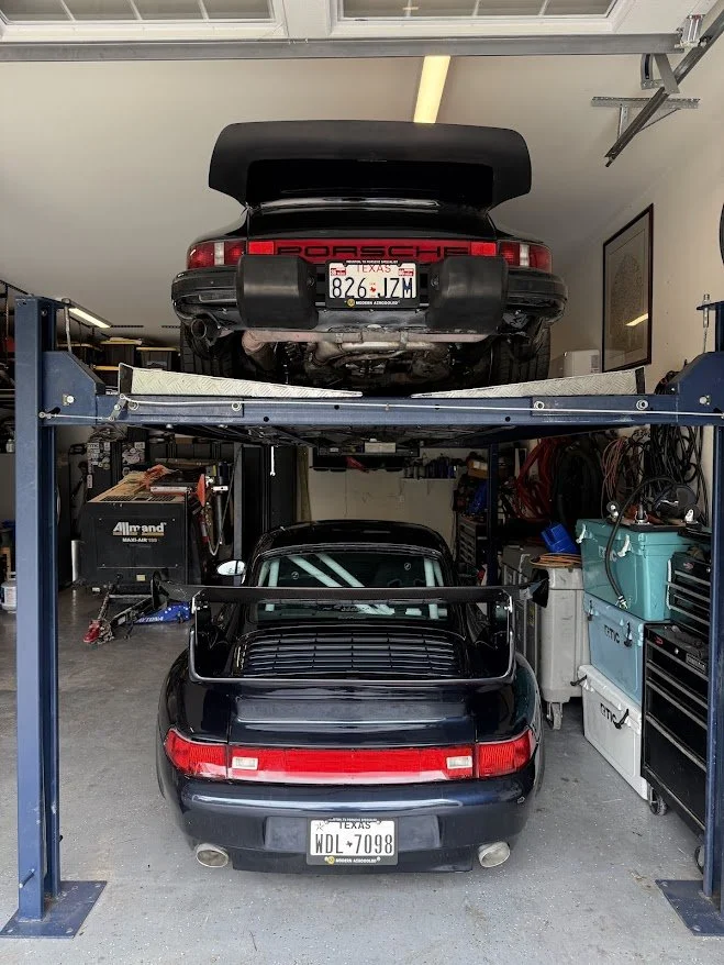 Black Porsche 911 with a large rear wing, parked inside a garage, viewed from the back. Another black Porsche 911 is elevated on a car lift above the first car.