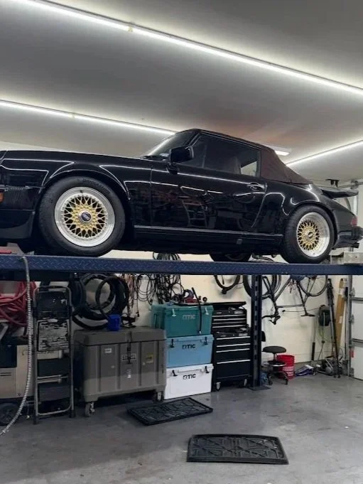 A black sports car with gold wheels is elevated on a lift in a garage, with tools and equipment underneath.