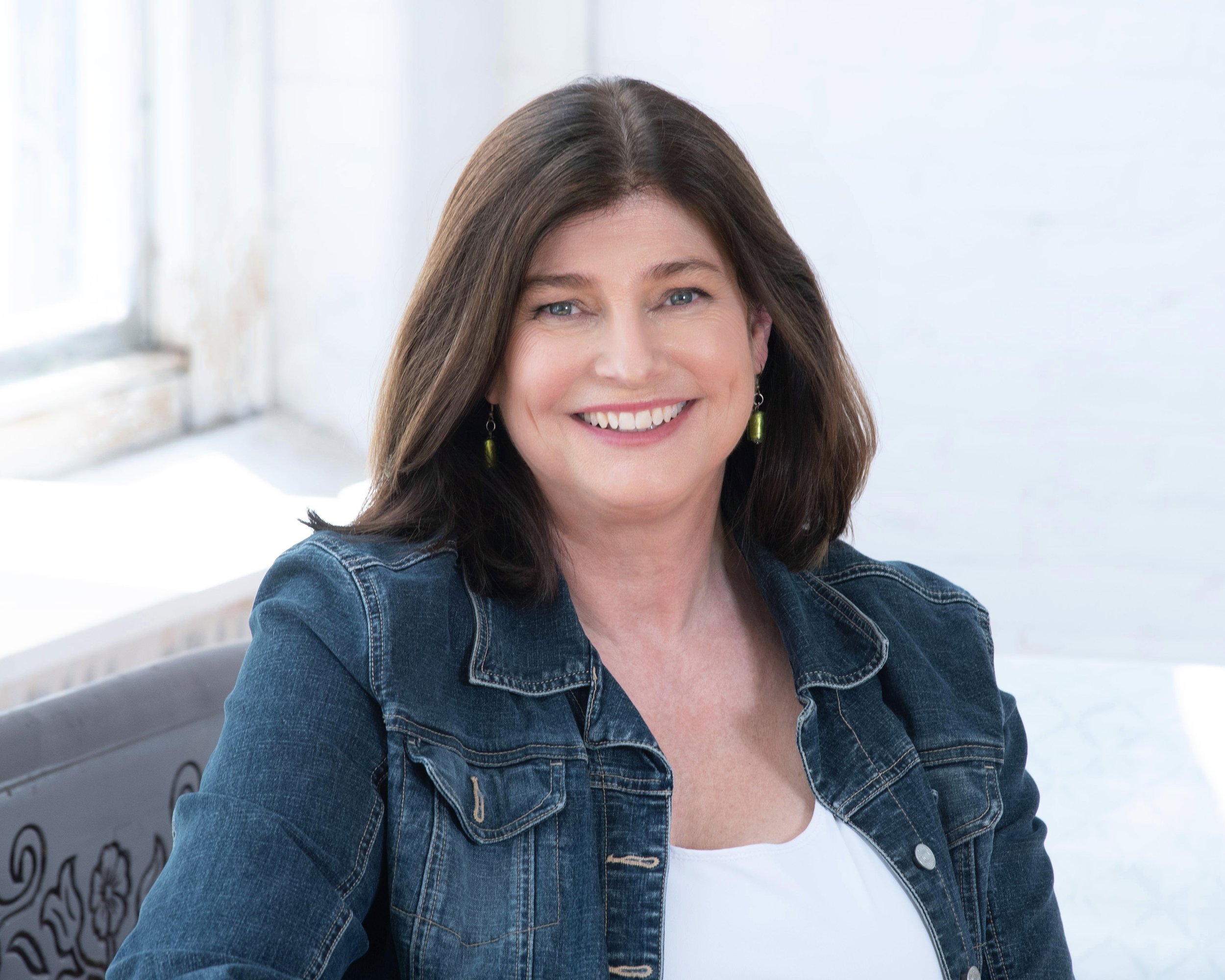 Smiling woman with brown hair and earrings sitting on a grey chair near a window with lace curtains, wearing a denim jacket and white top.