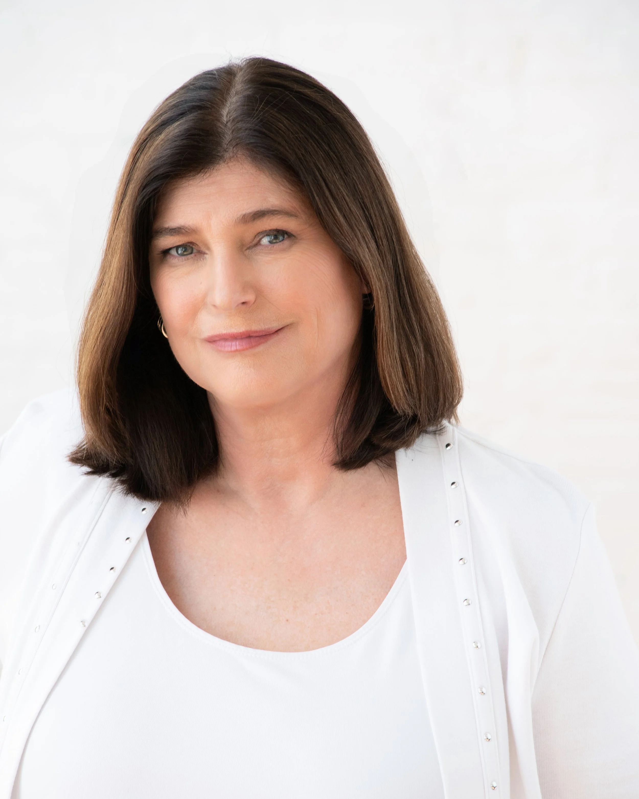 Close-up of a woman with shoulder-length brown hair, blue eyes, wearing a white top with pearl-like buttons, smiling softly against a white background.