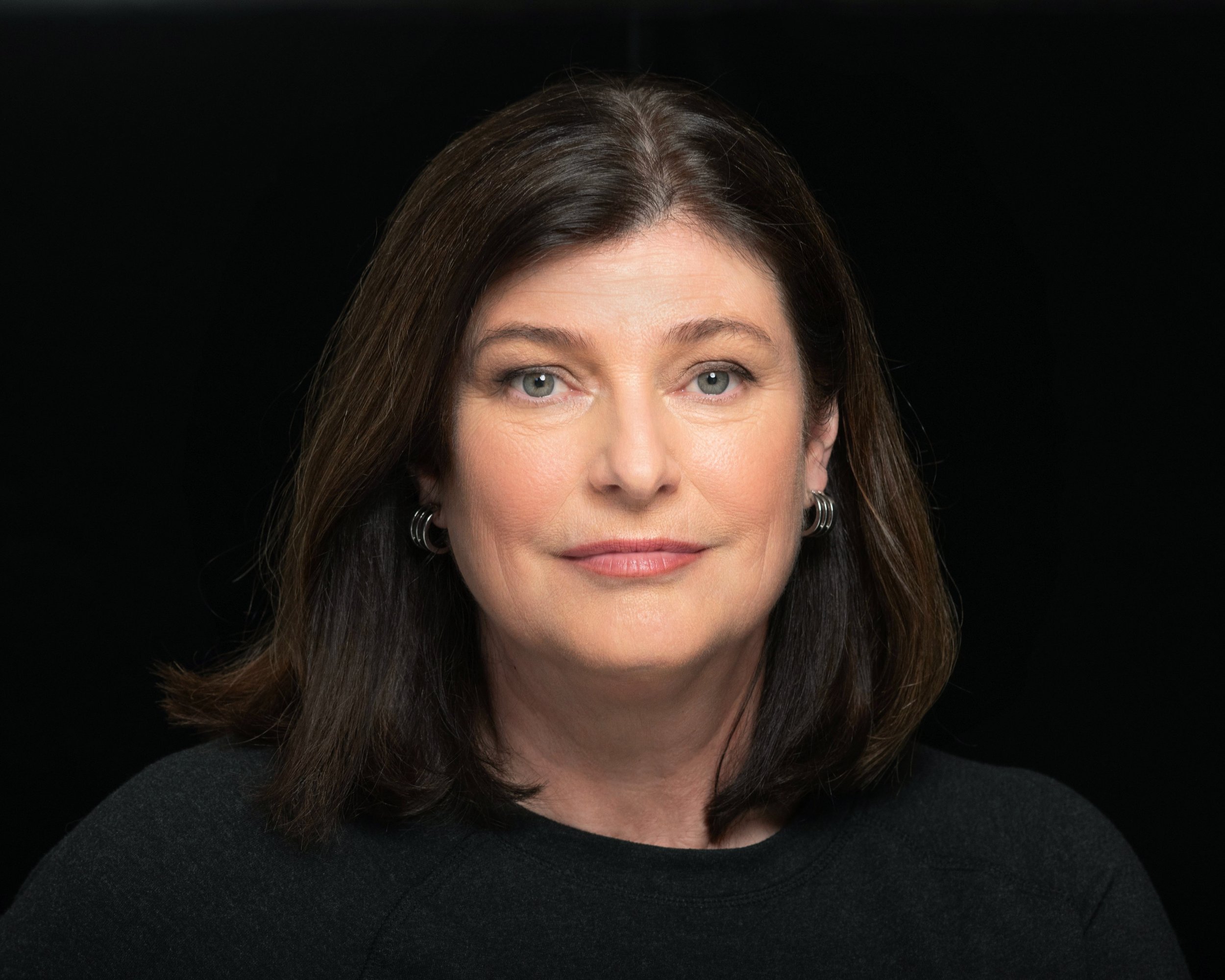 Close-up portrait of a middle-aged woman with shoulder-length brown hair, blue eyes, and earrings, against a black background.
