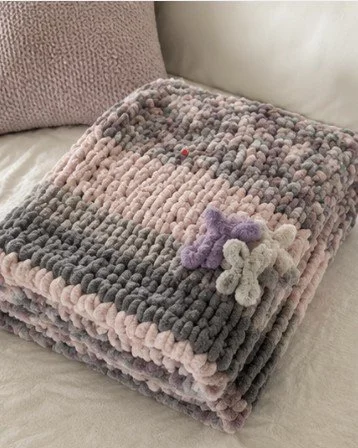 A folded multi-colored, textured blanket with a plush purple and white teddy bear toy placed on top, resting on a light-colored sofa with a purple pillow in the background.