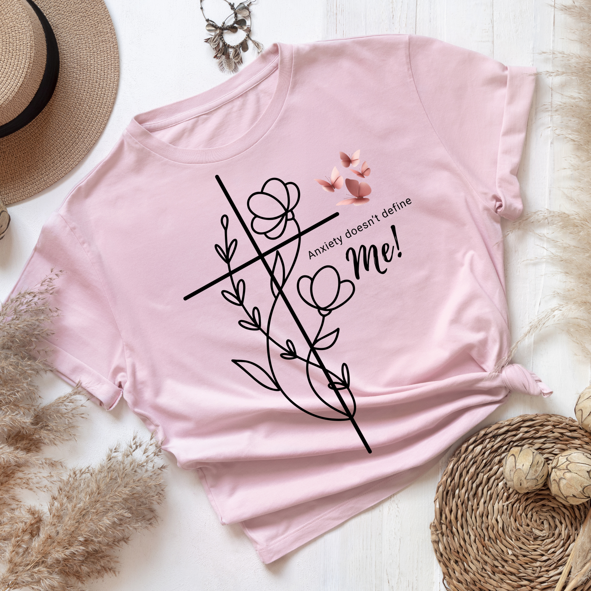 A pink T-shirt with black text and a decorative graphic, lying on a white wooden surface surrounded by a straw hat, feathered necklace, and woven coasters.