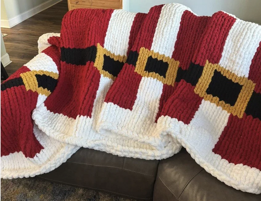 A Christmas-themed blanket with a Santa face design, draped over a dark brown couch.