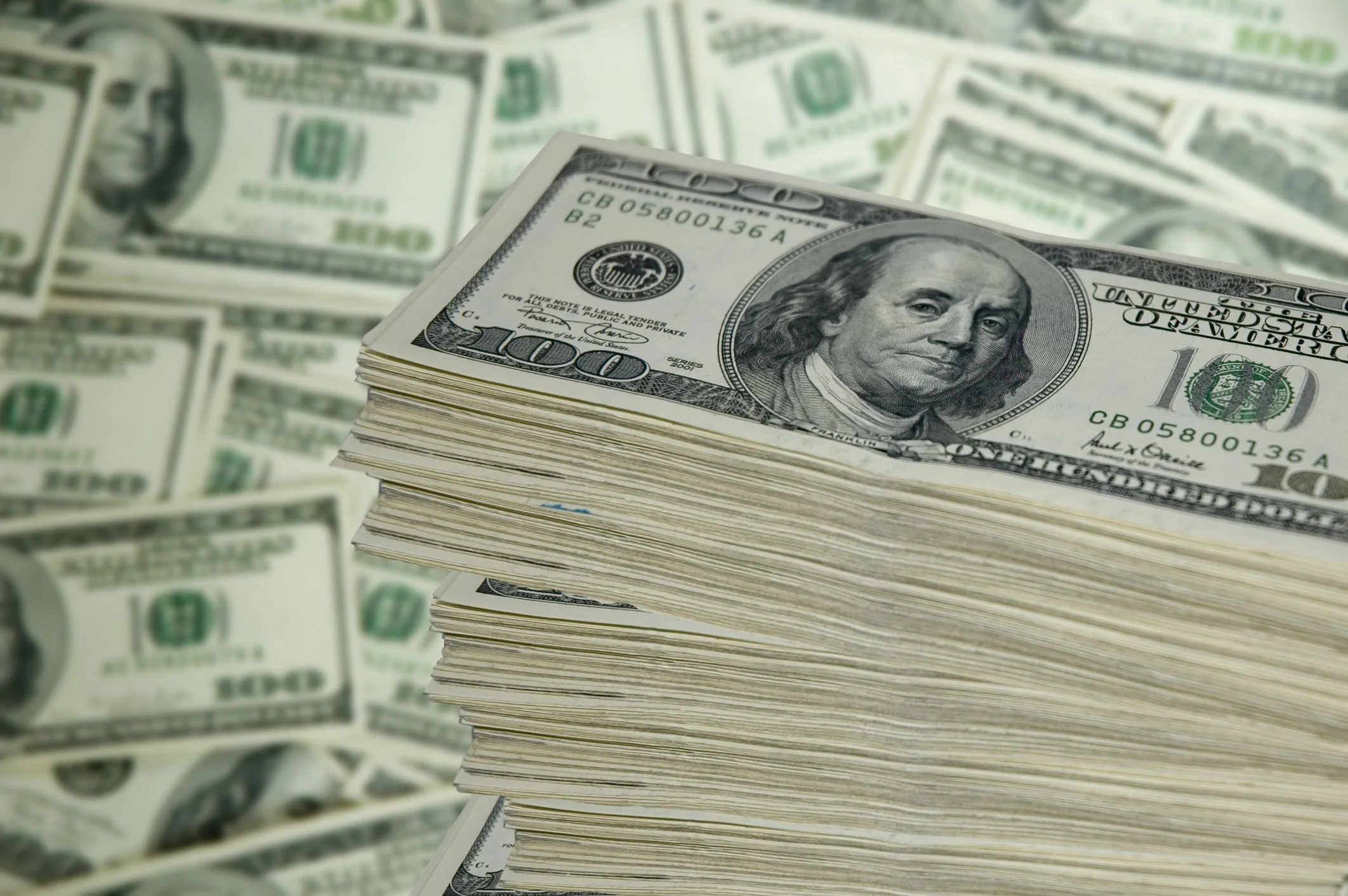 Close-up of a stack of 100-dollar bills in the foreground, with additional scattered 100-dollar bills in the background.