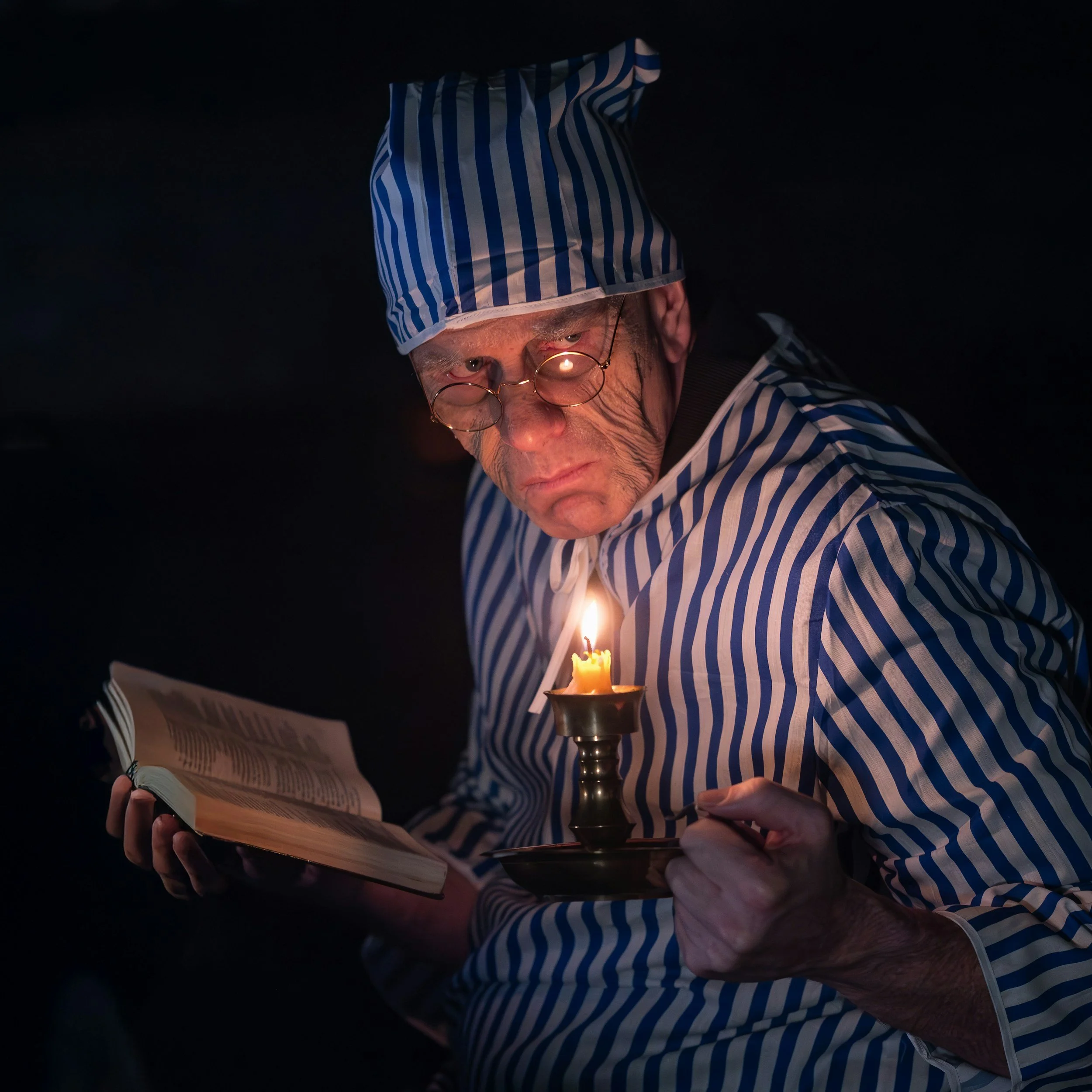 Close-up of an elderly man wearing striped pajamas and a striped nightcap, reading a book in low light with a candle in a holder in front of him.