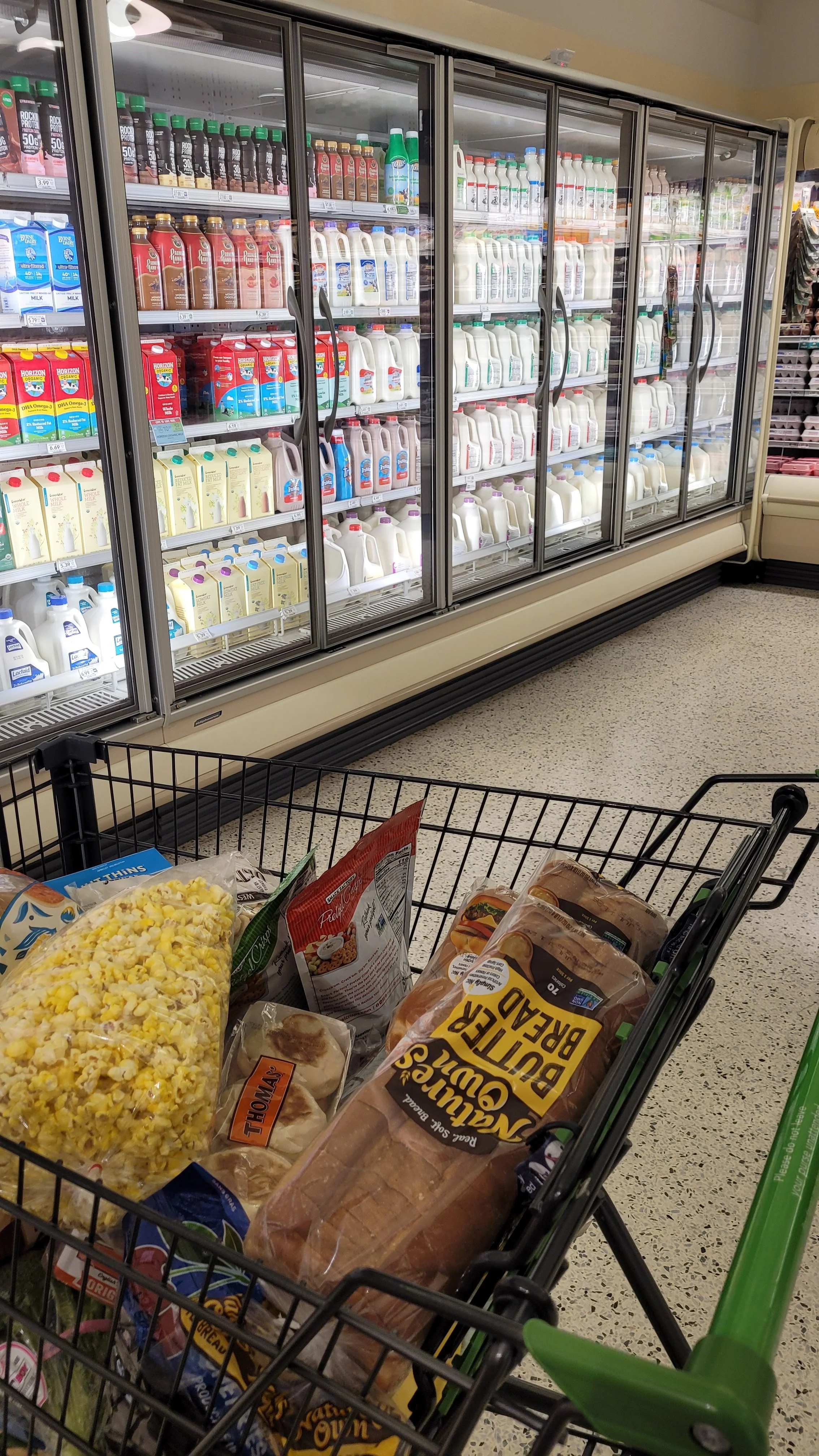 Grocery store cooler filled with bottles of milk, juice, and beverages. In the foreground, a shopping cart contains popcorn, bread, and other grocery items.