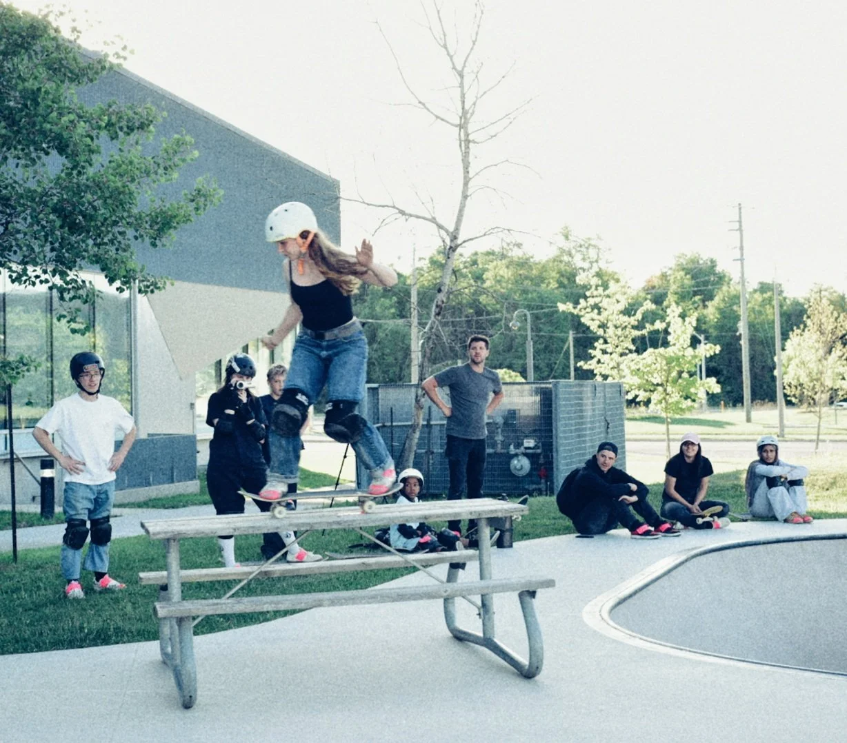 A girl wearing a white helmet, black tank top, and denim shorts skateboarding on a ramp with onlookers watching nearby at a skate park.