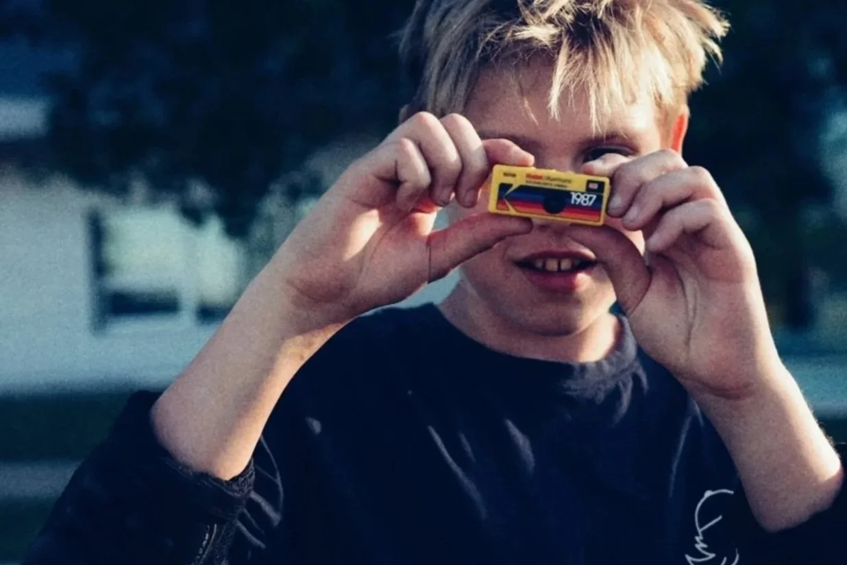 A young boy with blonde hair smiling while holding a small toy camera to his face, outdoors with trees in the background.
