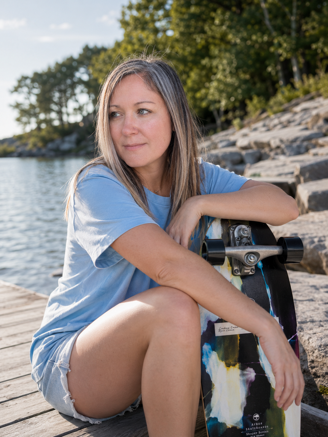 A woman sitting on a wooden dock by a lake, holding a skateboarding deck, with trees and rocks in the background during daytime.