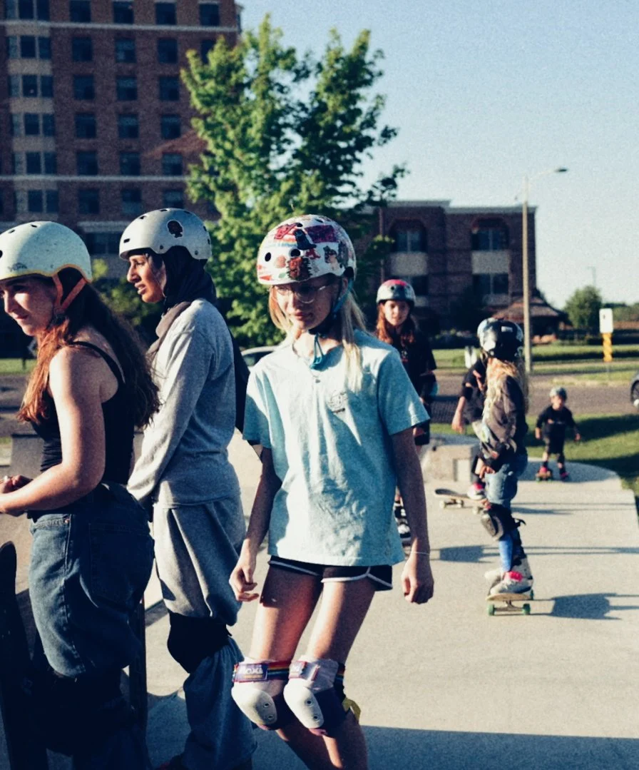 A group of girls roller skating outdoors on a sunny day, wearing helmets and protective gear, with trees and buildings in the background.