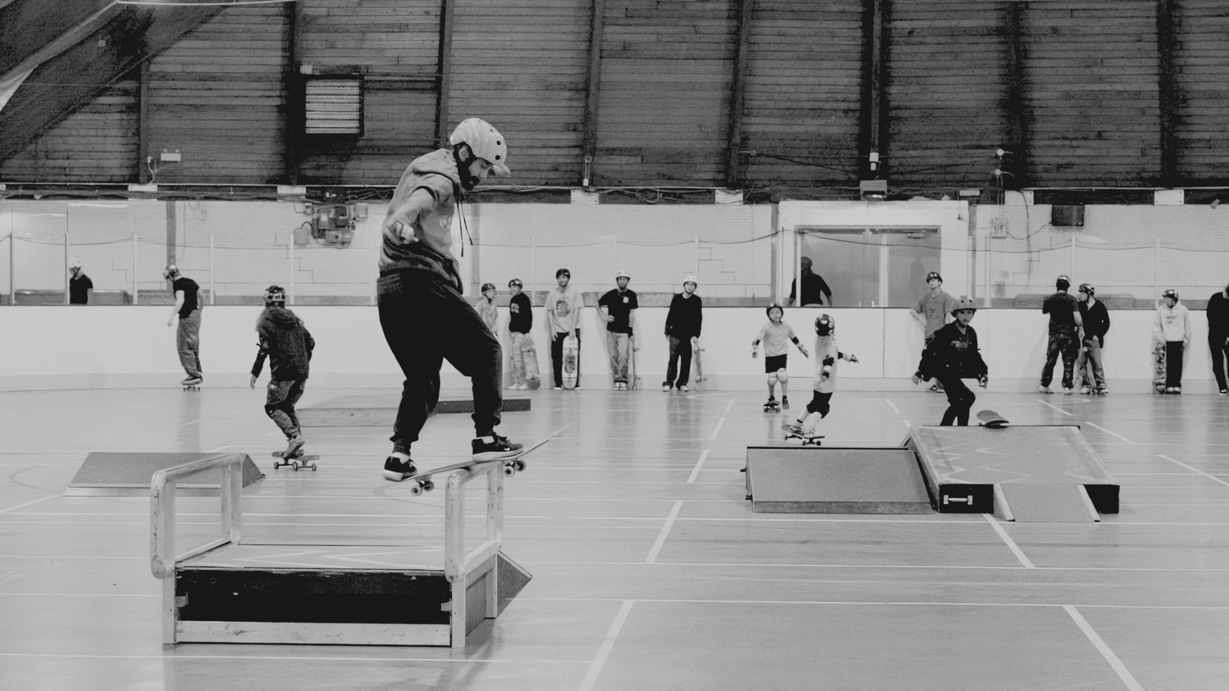 Skaters practicing at an indoor skate park with ramps and obstacles, with onlookers and coaches in the background.