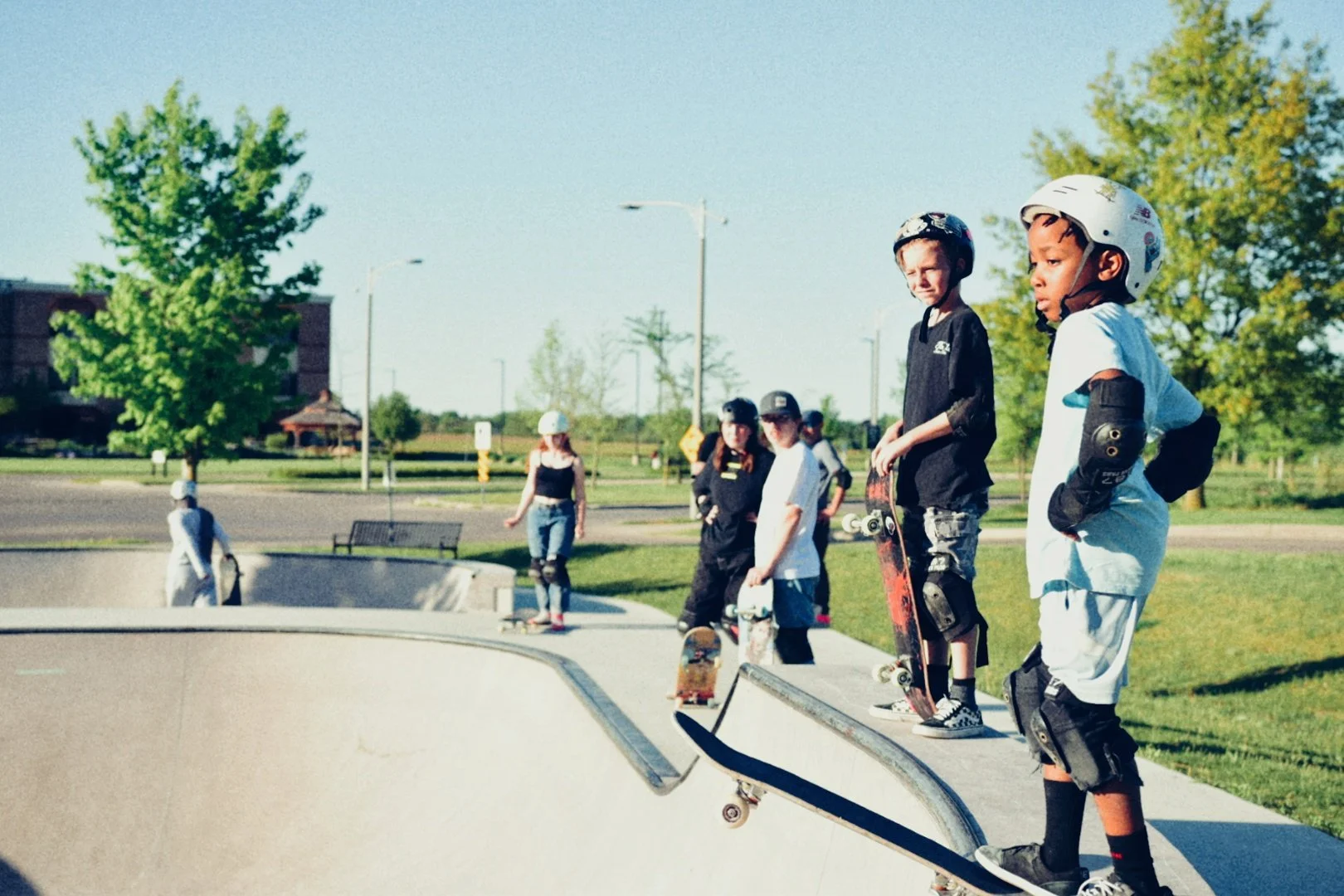Children wearing helmets and protective gear at a skate park, some holding skateboards, near a concrete skate bowl under a clear sky with trees and park benches in the background.