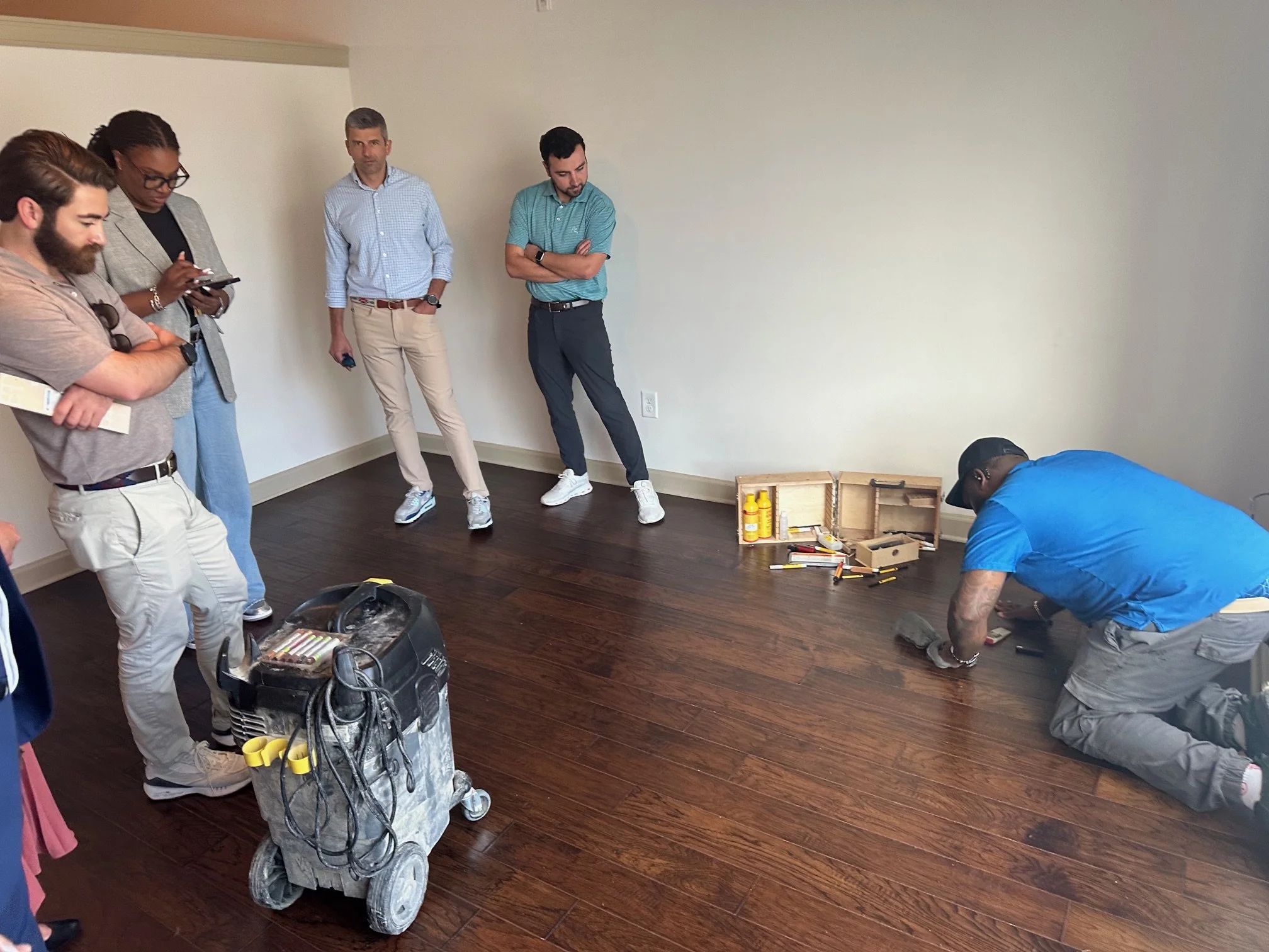 Group of five people standing and observing a man kneeling on the floor working on a small project. Supplies and tools are spread out on the floor and a toolbox in the foreground.