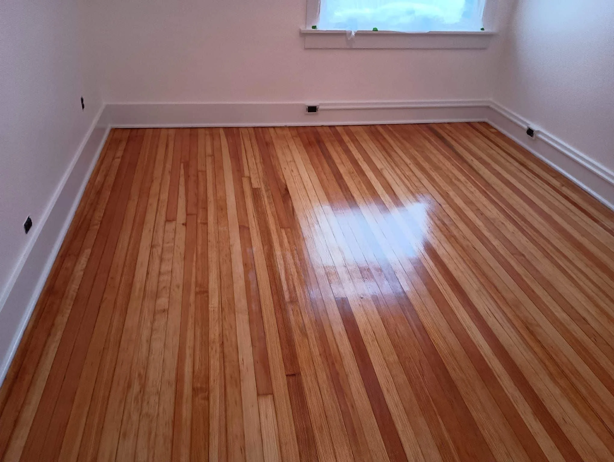Empty room with hardwood floor, white walls, a small window with sheer curtain, and electrical outlets on the walls.