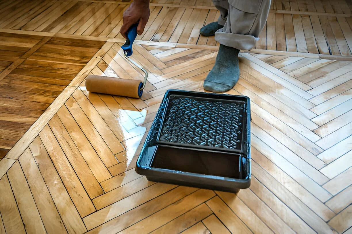 Person using a paint roller to apply clear finish on a wooden floor with a paint tray nearby.