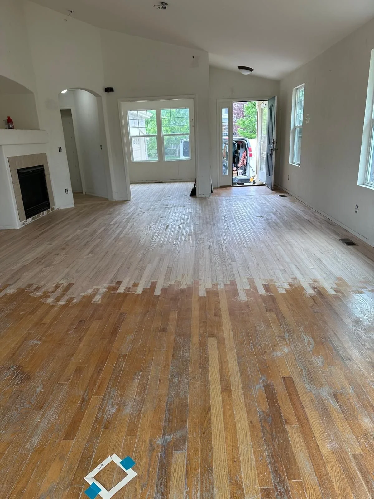 Living room with hardwood flooring in the process of refinishing or staining, with some areas still lighter and others darker, and a front door open to the outside.