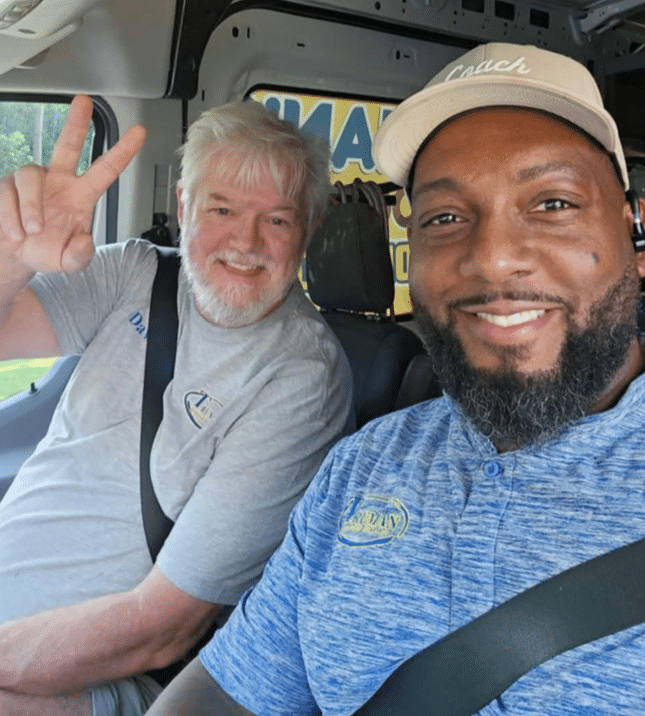 Two men sitting inside a vehicle, smiling at the camera. The man on the left has gray hair and a beard, wearing a gray T-shirt with a logo. The man on the right is wearing a blue shirt, a baseball cap, and has a beard. The background shows part of a sign that reads 'MAY' and some windows.