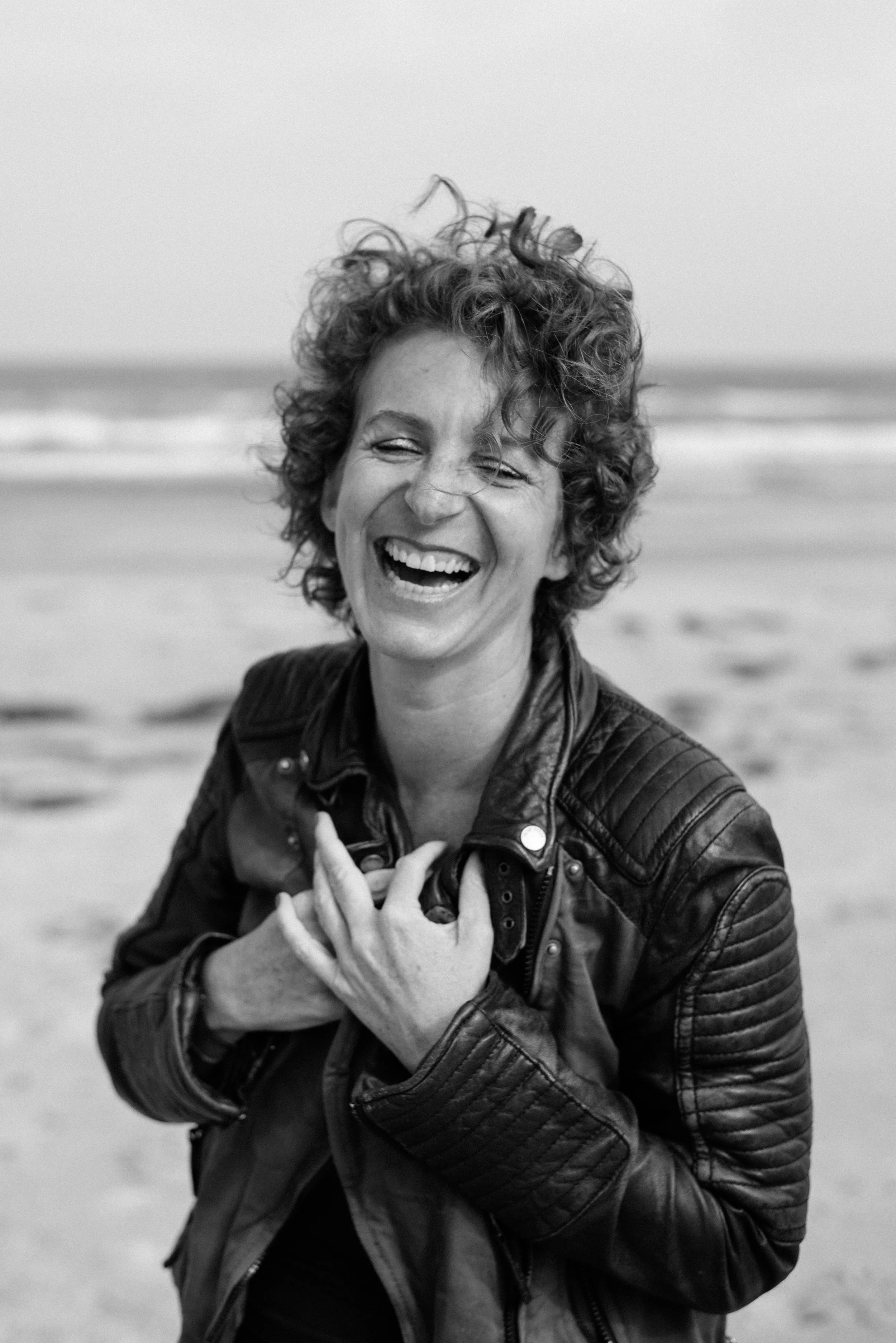 Schwarz-Weiß Fineart Fotografie einer Frau mit Locken und Lachen, in Lederjacke und am Strand mit Meer im Hintergrund