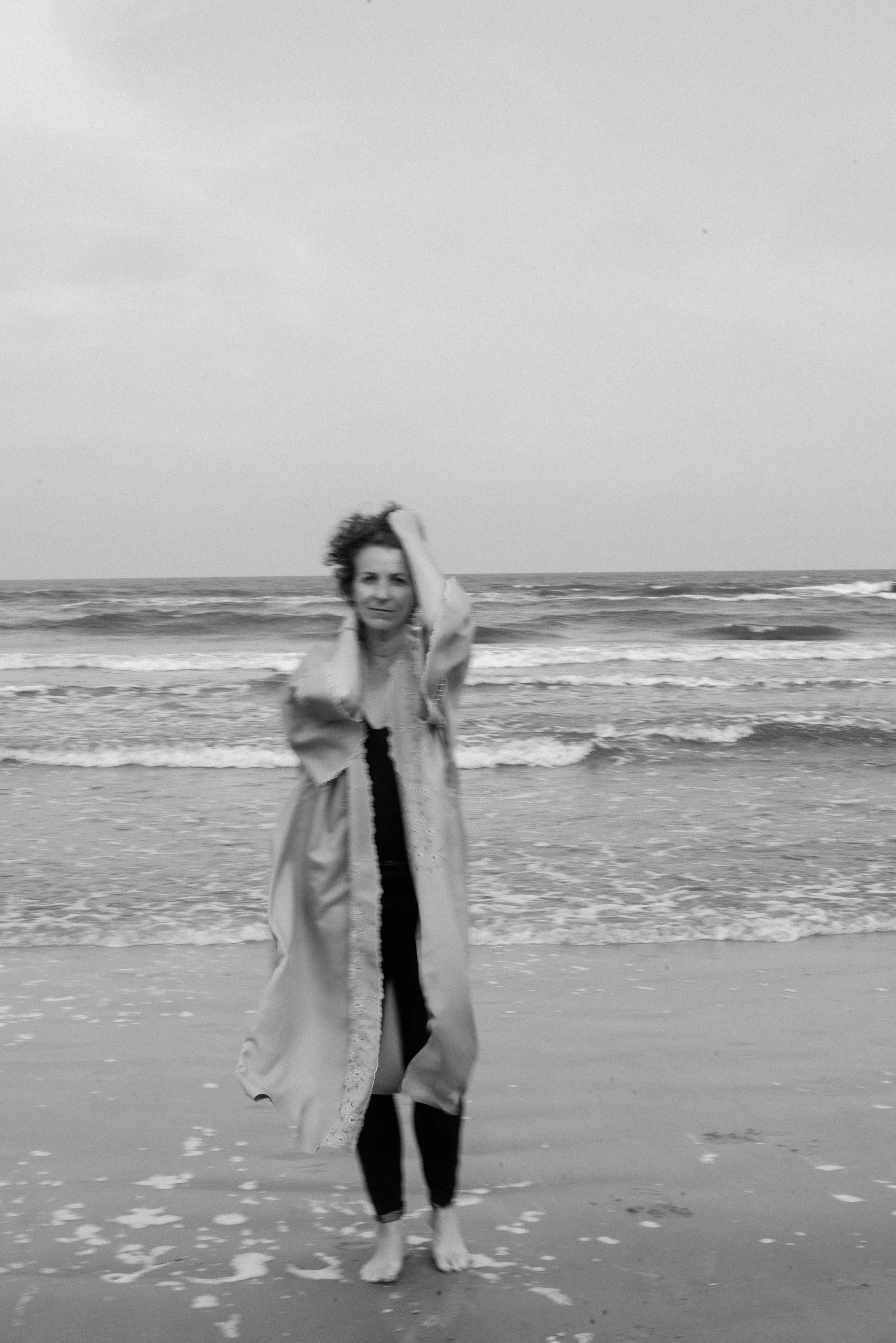 Eine Frau steht am Strand mit den Füßen im Meer, ein vom Winde verwehter Umhang, Meeresbrandung
