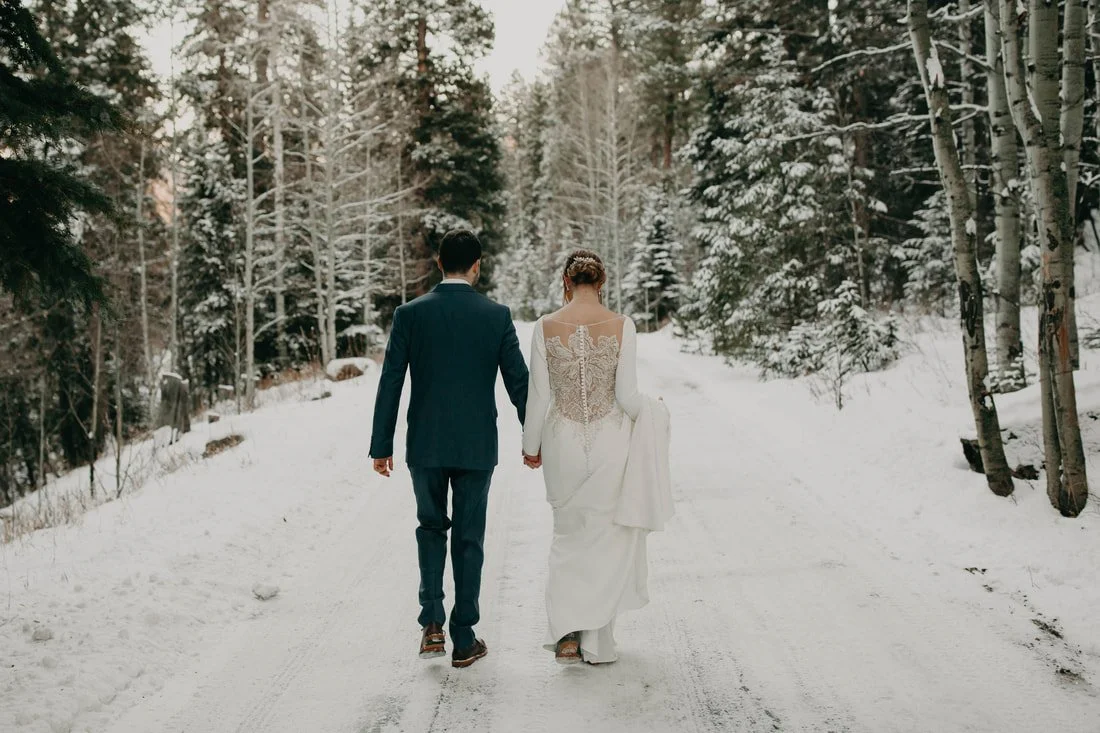 A bride and groom holding hands and walking in a snowy walkway, with snow-covered trees on both sides.
