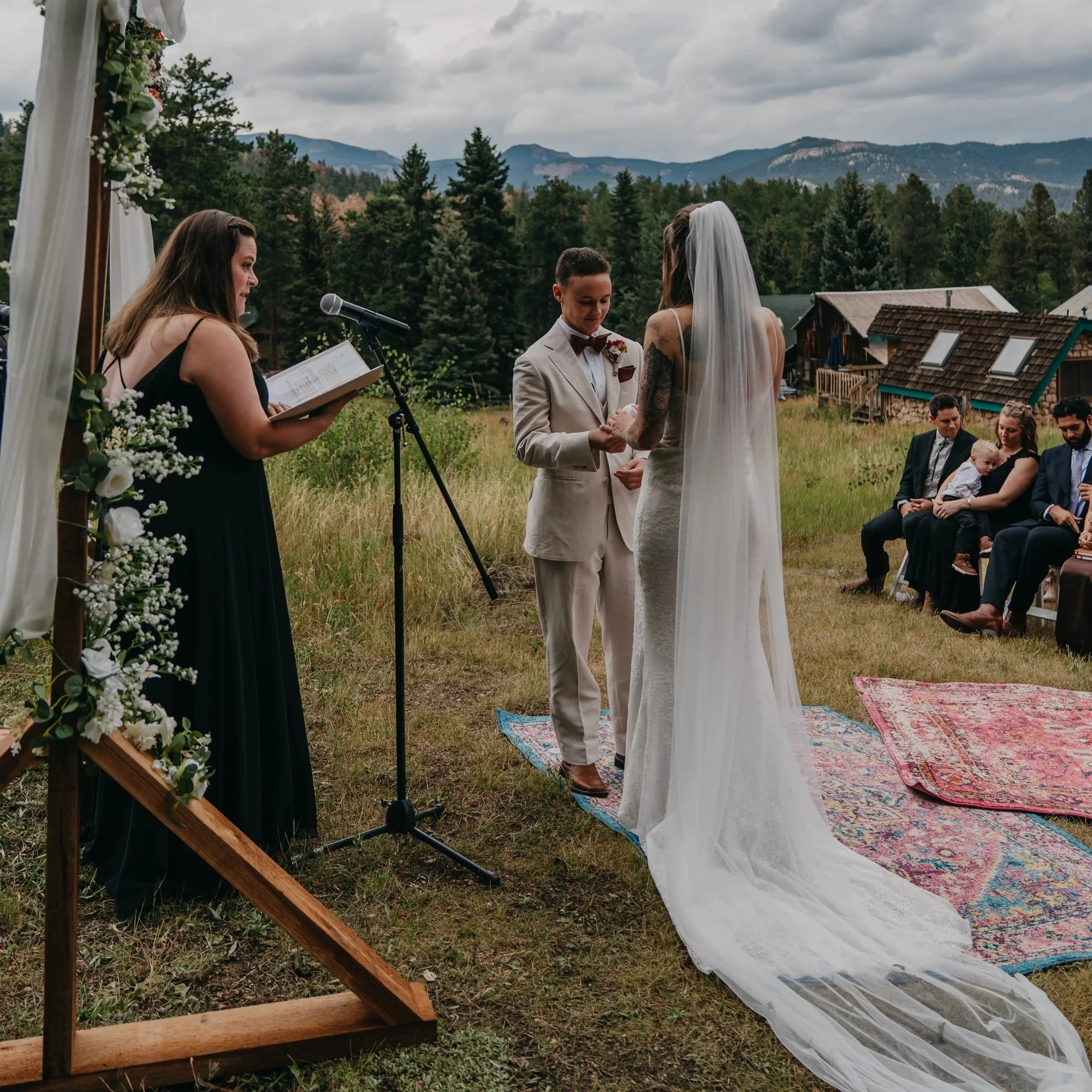 A wedding ceremony taking place outdoors on a grassy field with mountains and a cloudy sky in the background. The bride and groom are facing each other, holding hands, with guests seated nearby. An officiant is reading from a book.