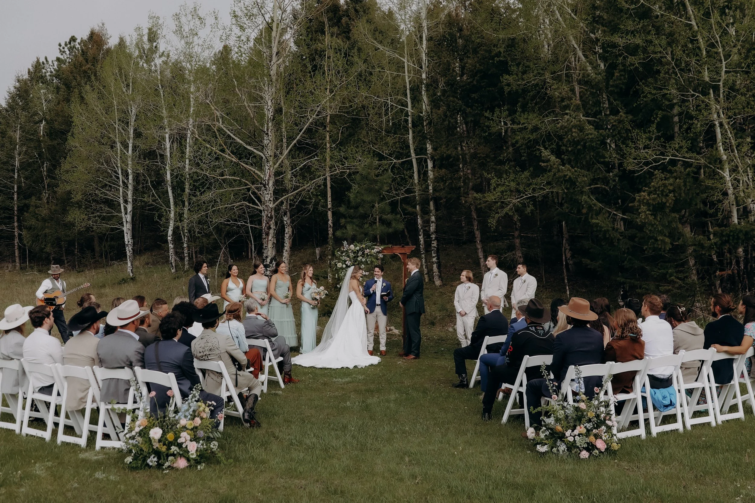 Outdoor wedding ceremony with bride and groom exchanging vows, surrounded by bridesmaids and groomsmen, audience seated in white chairs, officiant officiating, musician playing guitar, forested background.