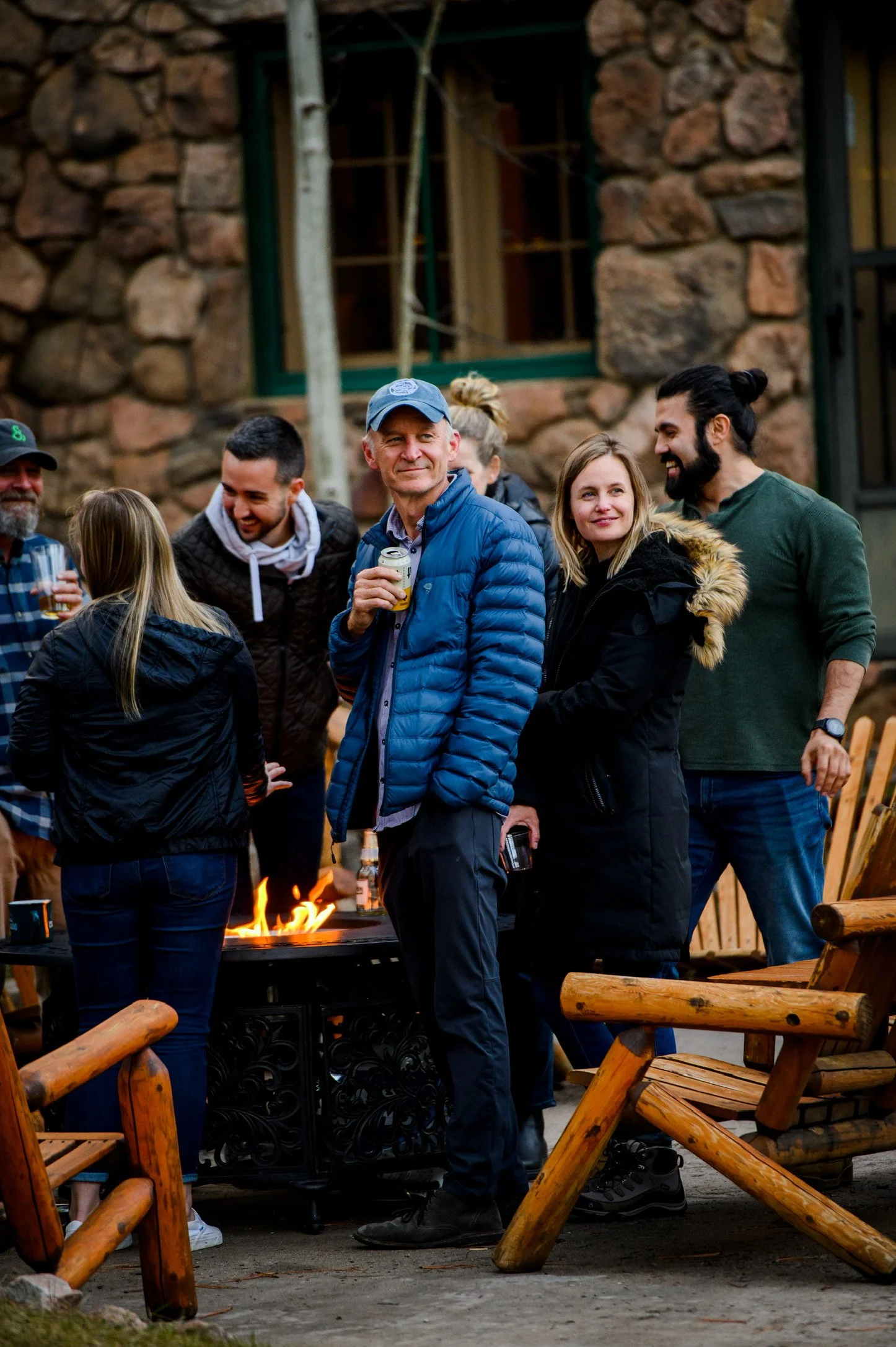 A group of people socializing outdoors near a fire pit with a rustic stone building in the background. They are smiling and holding drinks, enjoying a casual gathering.